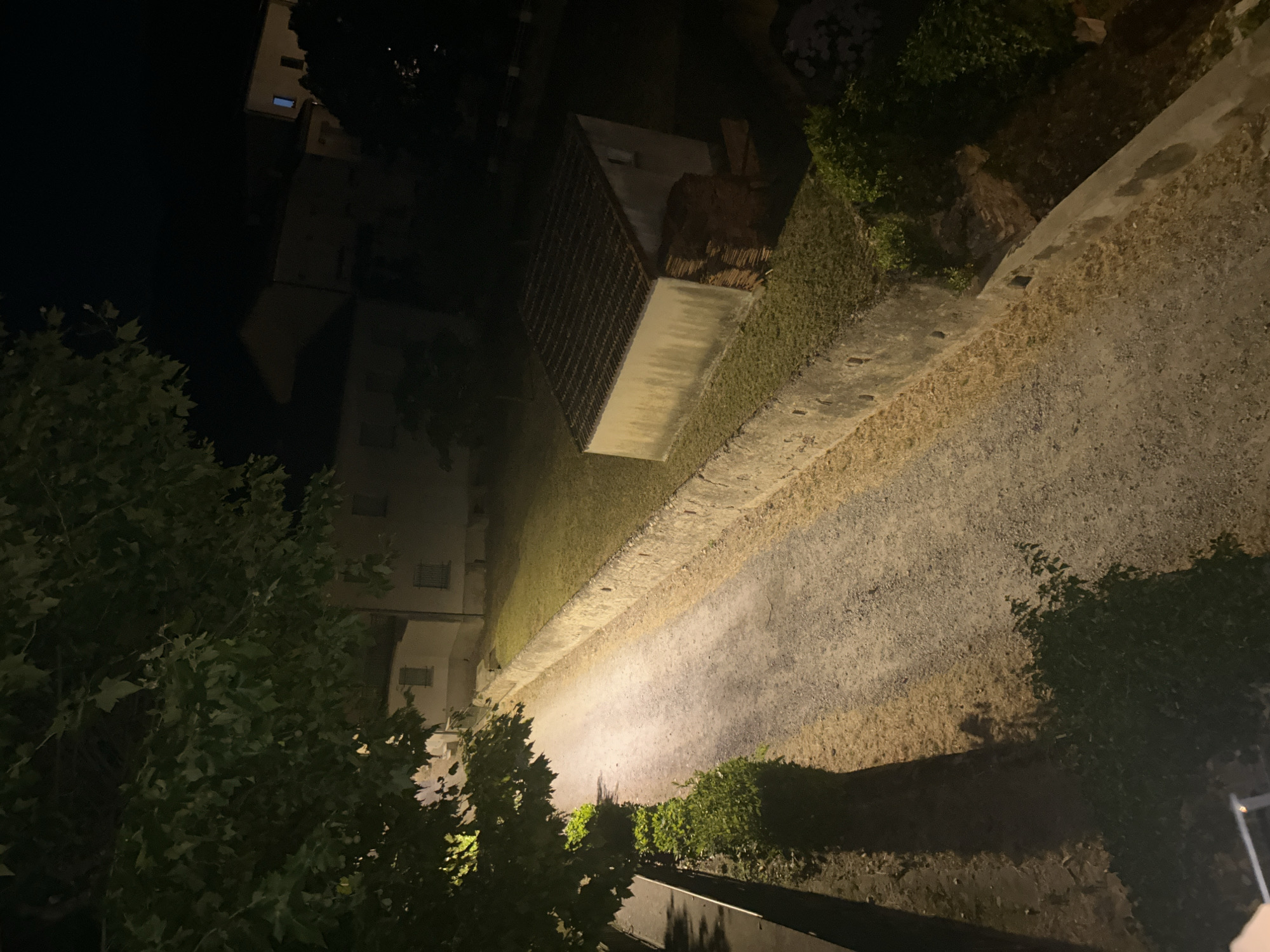 This is a night-time, outdoor, high-angle shot of a path or road illuminated by a light source, likely a streetlamp, casting long shadows. The path is bordered by a grassy area on one side and a low wall on the other. To the left of the path, a building with visible windows is partially obscured by dark green foliage. Further down the path, a large, rectangular structure with a grated top is visible, set into the grass. The scene is dark, with the light source creating a stark contrast between illuminated areas and deep shadows. The overall impression is one of quiet, possibly deserted, nighttime ambiance. There is no visible text in the image.