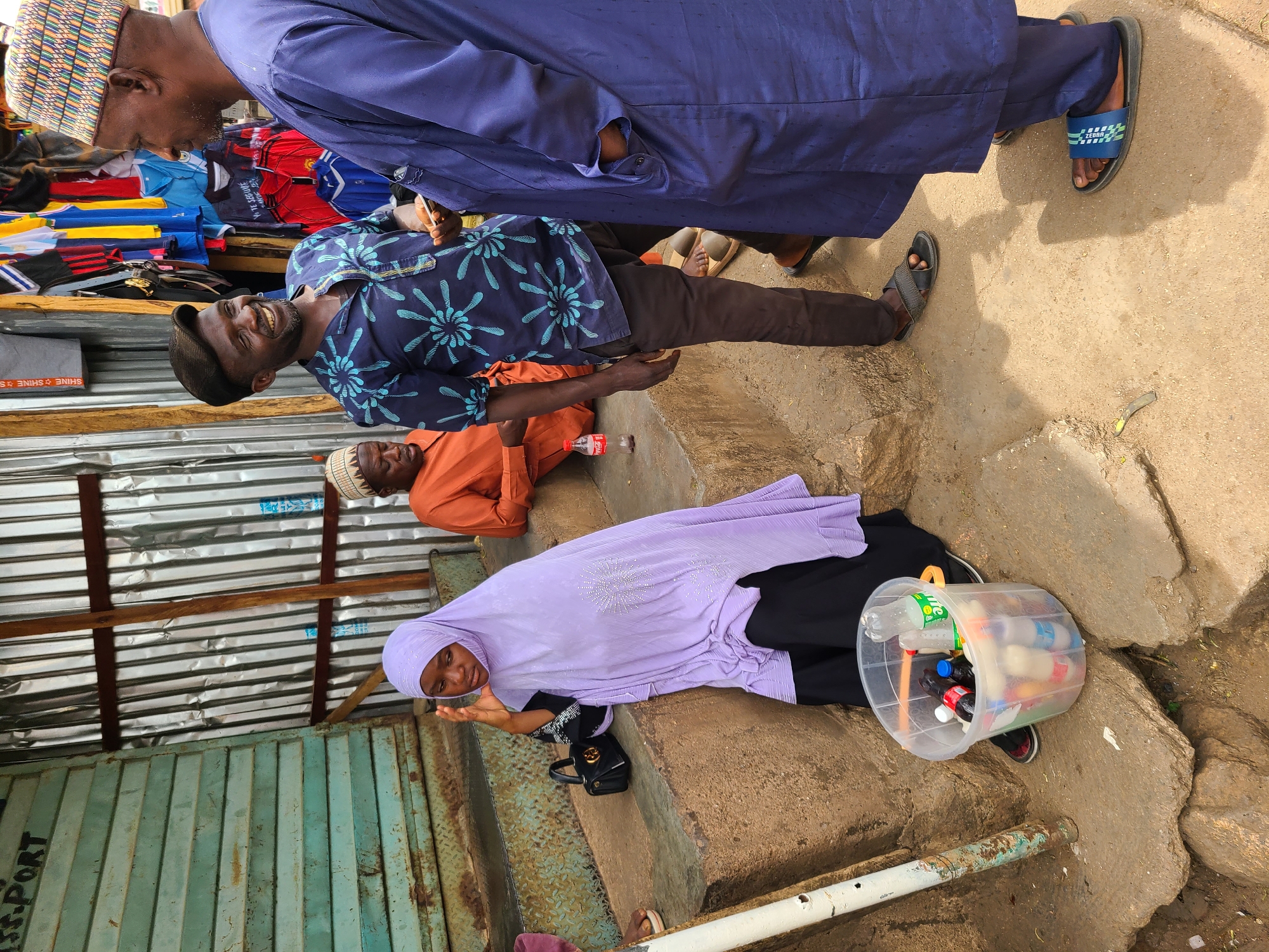 In Jos, Nigeria, a woman wearing a lilac headscarf and black clothing sits on concrete steps near a corrugated metal wall, with a clear plastic bucket filled with bottles of various beverages positioned next to her. A man in an orange shirt lies on the step above her, while two standing men, one in a blue floral shirt and one in a blue robe, are positioned above. The backdrop includes a metal structure and the ground is a mix of concrete and bare earth.