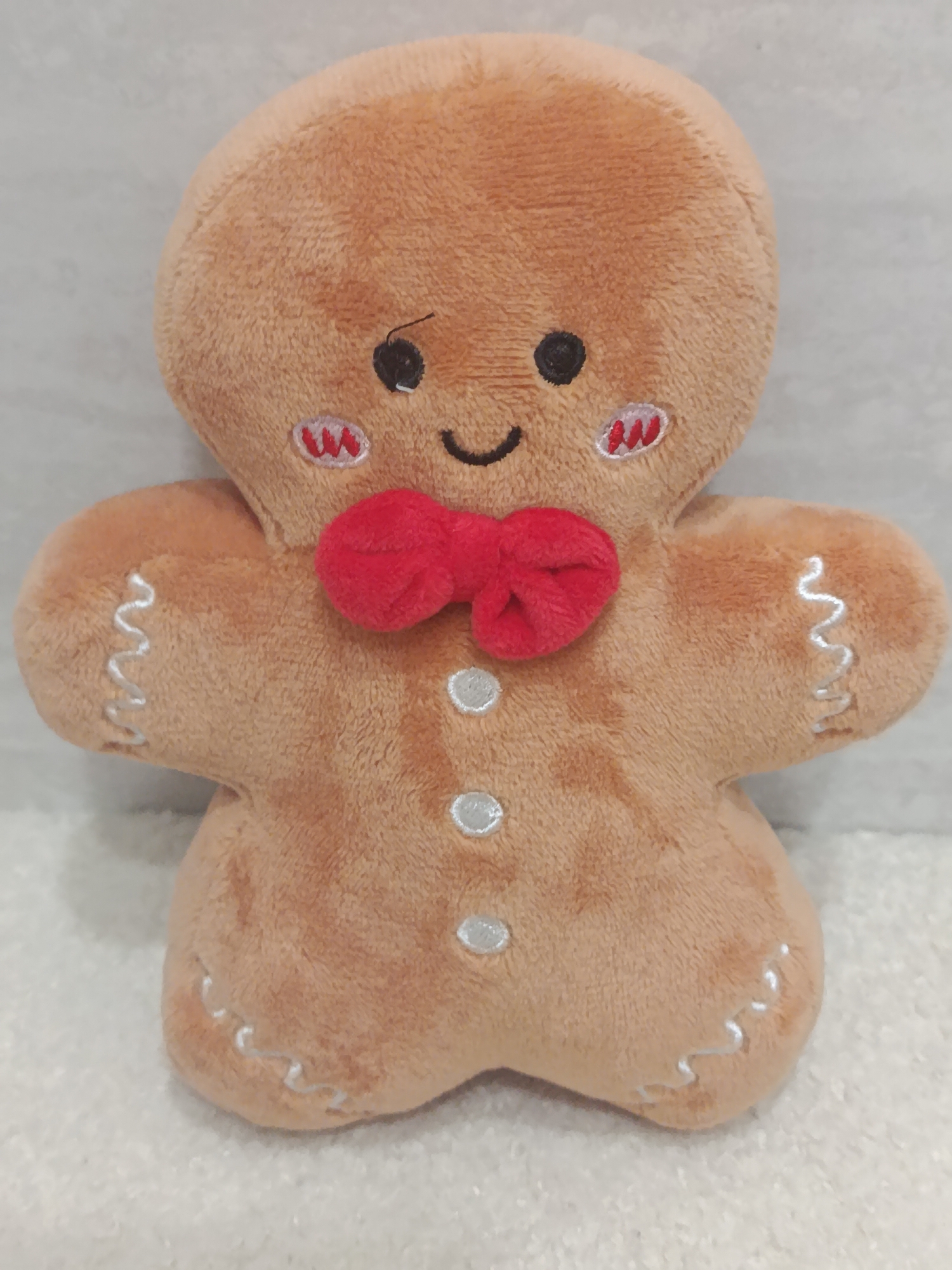 This is a close-up, eye-level, studio shot of a plush gingerbread man toy. The toy is a light brown color with embroidered features. It has black button eyes, a black embroidered smile, and rosy cheeks with red embroidered accents. A small red bow tie is attached at the neck. Three white buttons are sewn down the center of the torso. The arms and legs have white embroidered zig-zag patterns. The background is a textured white surface, likely fabric, with a lighter gray, textured wall behind it. There is no indication of a specific location in Chișinău, Moldova.
