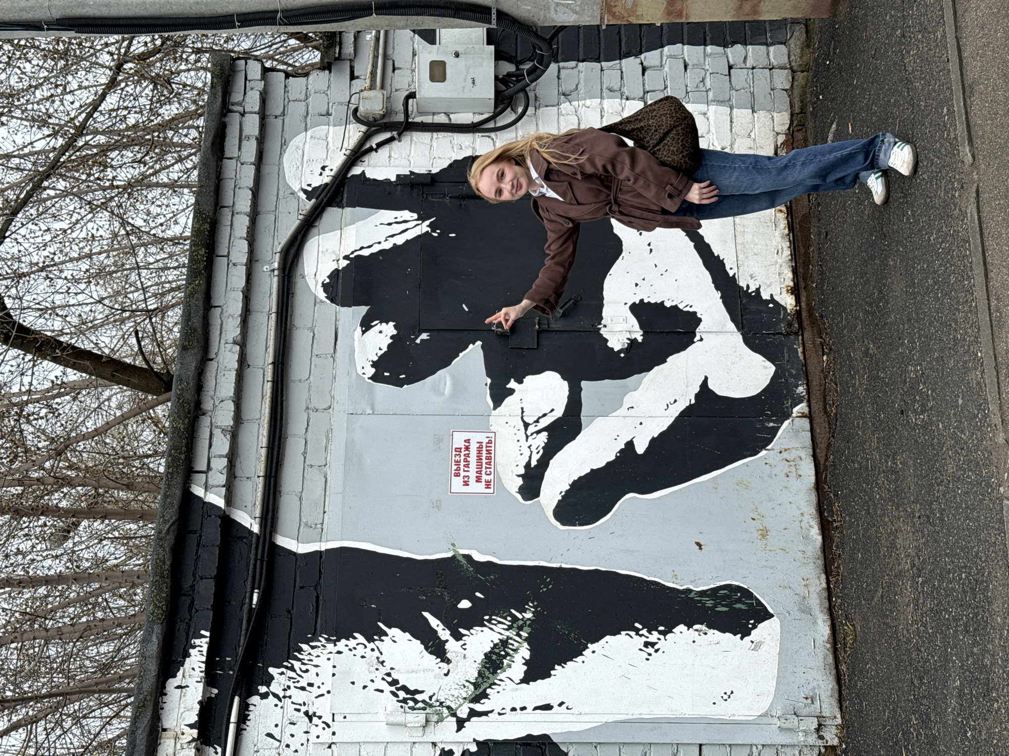 A photograph taken outdoors in the city of Perm, Russia, shows a young woman standing next to a garage door painted with a black and white mural depicting two hands nearly touching. The mural appears to be a representation of the “Creation of Adam” by Michelangelo. The woman wears a brown jacket, blue jeans, and white sneakers. She points towards the space between the hands in the mural. A sign is affixed to the garage door, bearing text written in Russian. A leafless tree is visible to the left of the garage door, and pavement is visible to the right. Electrical wiring and a utility box are present above the garage door.