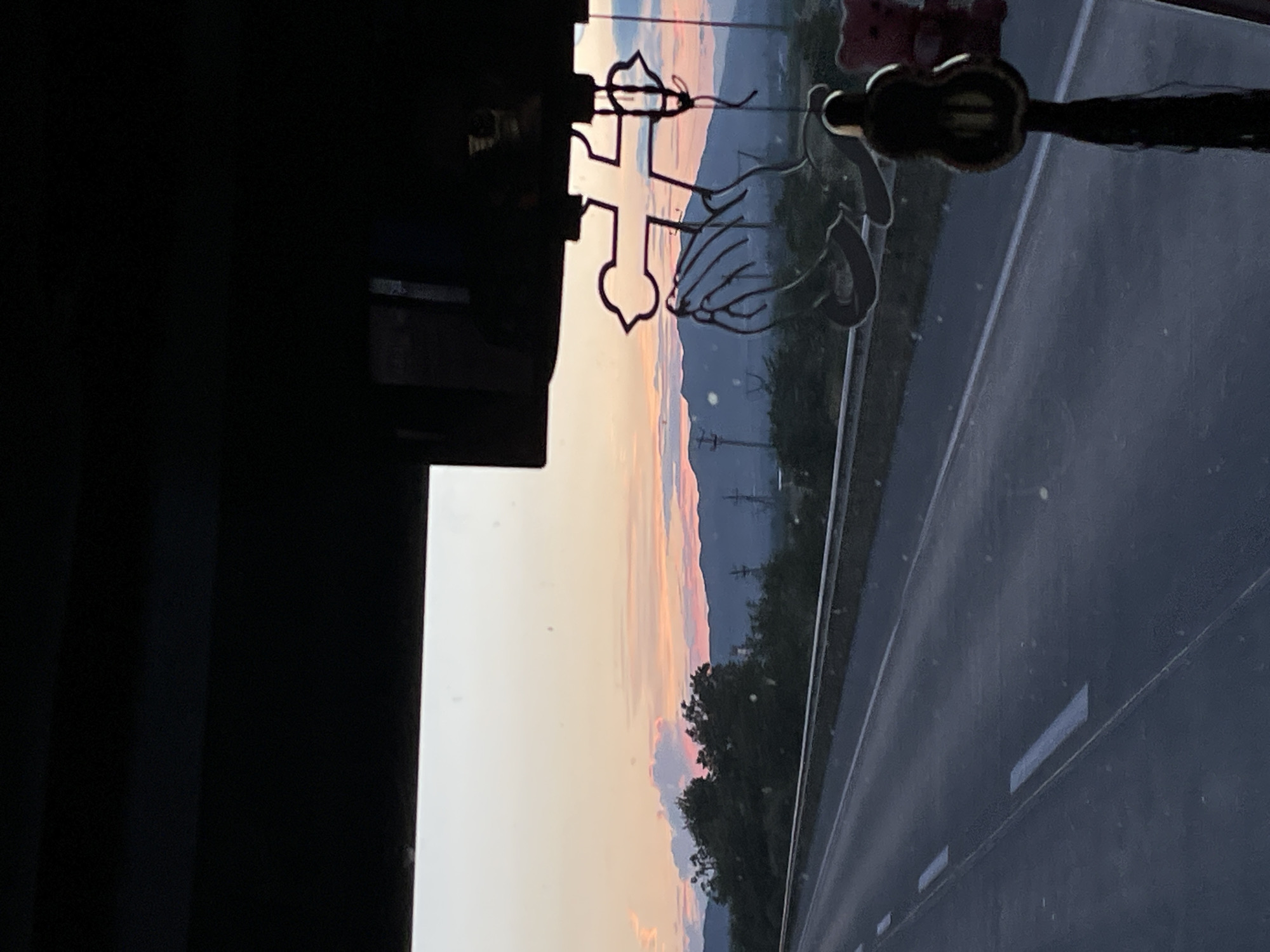 The image shows a view from inside a vehicle, looking out at a highway at sunset. The sky displays soft hues of pink, orange, and yellow. In the foreground, on the right side of the windshield, a religious ornament featuring praying hands and a cross is visible. Below this ornament, a dark, teardrop-shaped charm with a tassel hangs down. A small pink teddy bear is attached to the right side of the ornament. The highway stretches into the distance, with markings on the asphalt. Beyond the highway, there are trees and hills, with power lines visible in the middle ground. The overall scene suggests a journey through the Georgian countryside near Dzveli Kanda as the day ends.