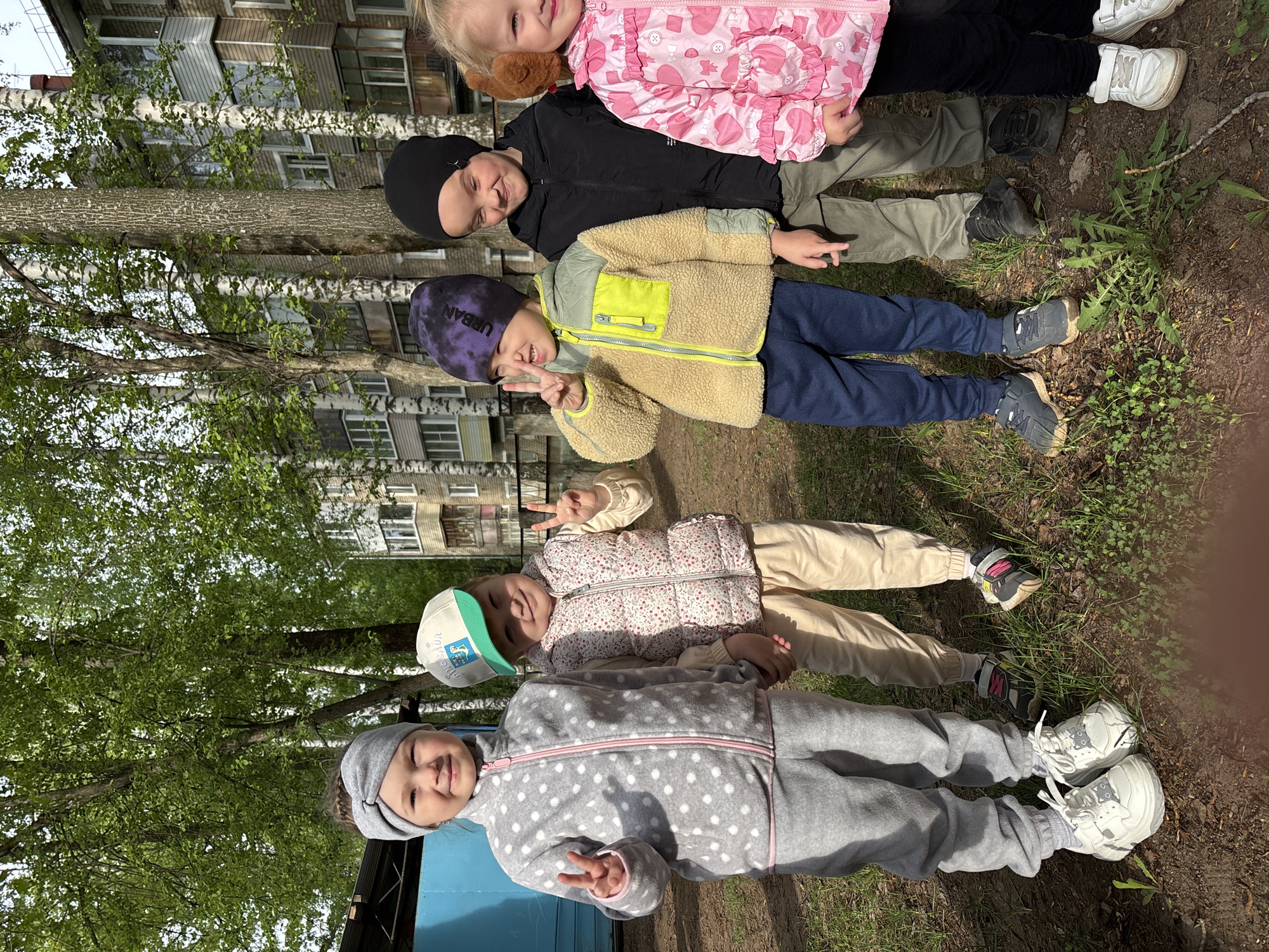 A line of five children are lying head-to-toe on a patch of grass near a blue metal structure. The children are dressed in outerwear appropriate for cool weather. The setting appears to be outdoors, possibly a park or playground, with trees and a building visible in the background. Given that the location is Chaikovsky, Russia, the building is most likely a typical urban apartment complex found in many Russian cities. The first child, closest to the blue structure, is wearing a gray polka-dot jumpsuit with a gray headband and is making a peace sign with their hand. The second child is wearing a beige vest, a white hat, and light-colored pants and is also making a peace sign. The third child has on a purple and blue patterned beanie, a tan jacket with yellow accents, and dark blue pants. They are also displaying a peace sign. The fourth child is wearing a black jacket and a black beanie. The fifth child is wearing a pink patterned jacket. The background includes birch trees, suggesting a common landscape in Russia.