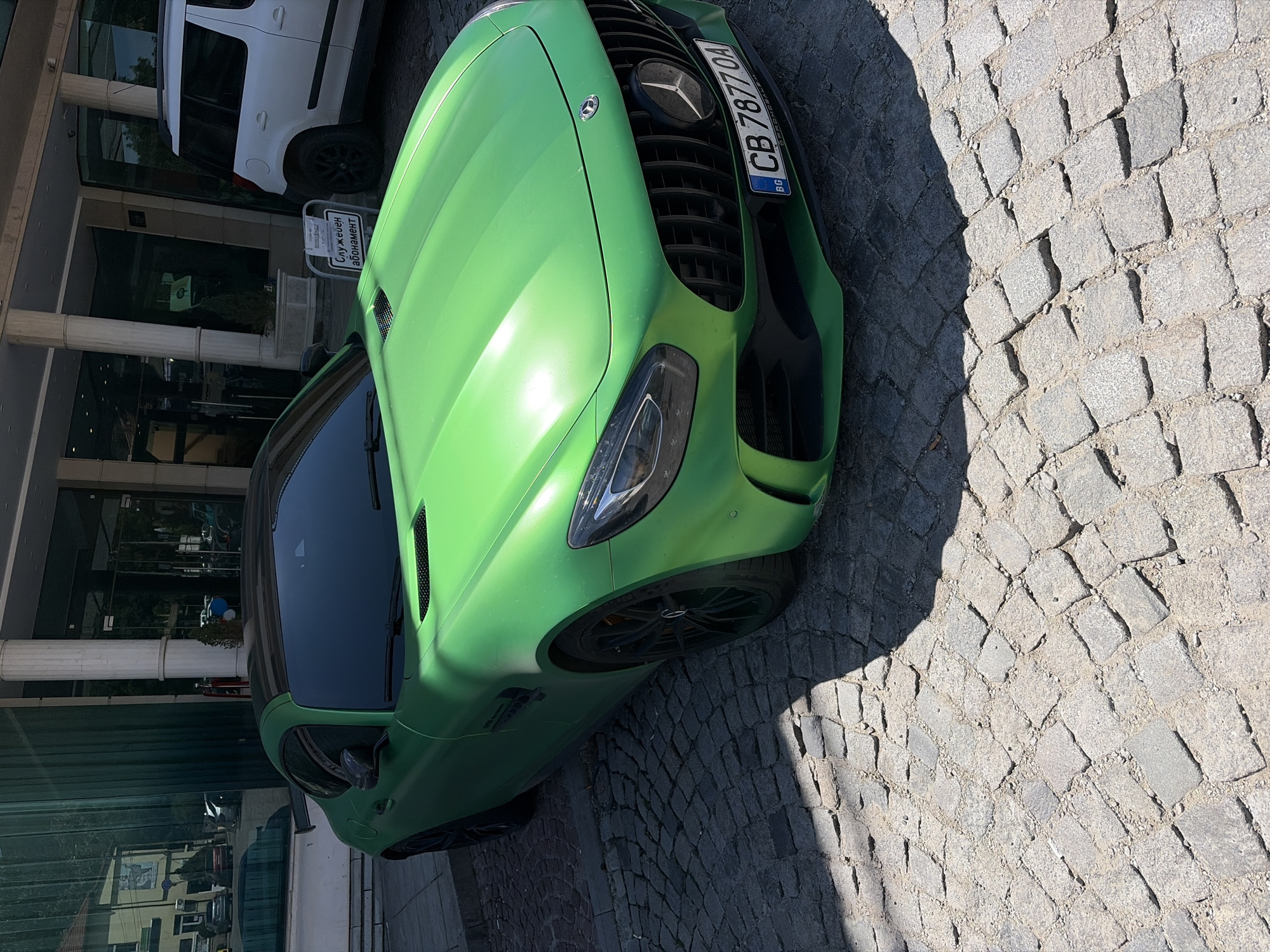 A vibrant green Mercedes-Benz AMG GT is parked on a cobblestone street in front of a building with large glass windows and white pillars. A white SUV is parked to the right of the sports car. A sign with Bulgarian text, likely indicating parking information, is visible between the two vehicles. The sky is clear and blue, suggesting a sunny day. The scene appears to be in an urban setting, possibly a hotel or a commercial building. The license plate of the Mercedes reads "CB 7877 OA".