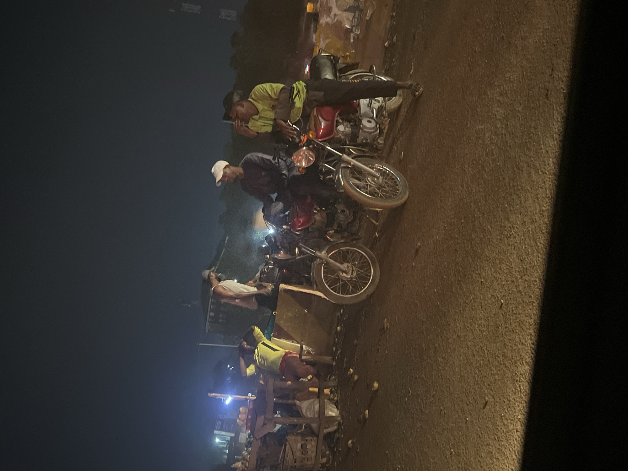 The image captures a street scene at night in what appears to be a commercial or market area. Several individuals are visible. Two men are sitting on red motorcycles parked on the side of a paved road. One man is wearing a light green shirt and the other a gray shirt and white cap. Down the road, another man in a white tank top is standing next to wooden stands with various items on them. One individual in yellow and red clothing is at the wooden stands. The environment includes street lighting, and in the background, there are buildings, suggesting an urban area. Based on the information provided, the location is the city of Abuja, Nigeria.