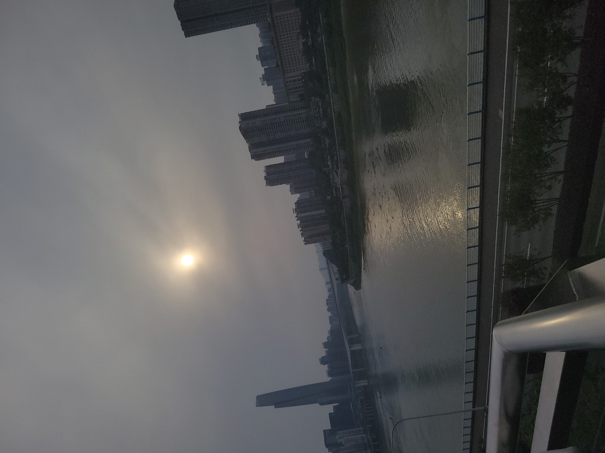 The image presents a cityscape in Yuzhong District, China, viewed from an elevated perspective, likely a bridge. The scene is characterized by overcast conditions, with the sun visible through a layer of haze or fog.
To the right, a river runs lengthwise, reflecting the dim light. The riverbank is lined with trees and a pedestrian walkway, bordered by a metal railing, part of which is prominently featured in the foreground.
On the left bank, a dense cluster of high-rise buildings dominates the skyline. The buildings vary in height and architectural style, representing the urban environment.
Overall, the image captures an atmospheric view of a Chinese city, emphasizing its urban landscape and riverfront location in Yuzhong District.