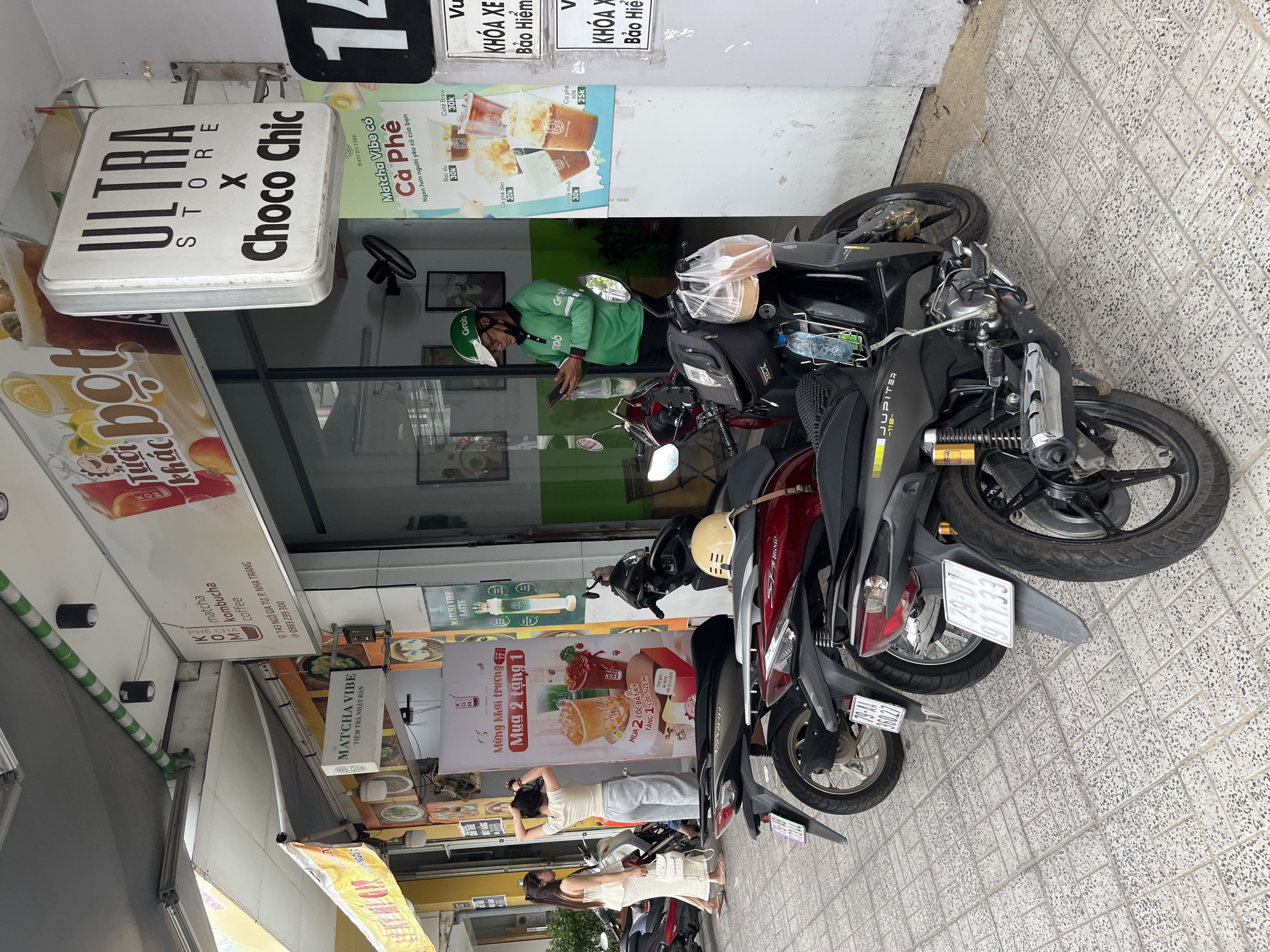 The image depicts a street scene in Nha Trang, Vietnam, likely outside a cafe or shop. In the foreground, several motorcycles are parked on a tiled sidewalk. A man wearing a green Grab uniform and helmet is standing next to a black motorcycle, looking at his phone and holding a plastic bag. Behind him, two women are standing outside a building with awnings. The building features signage for "ULTRA STORE X Choco Chic" and "Matcha Vibe". Various advertisements for drinks are also visible on the building and a large banner. The scene suggests a commercial area with food and beverage services.