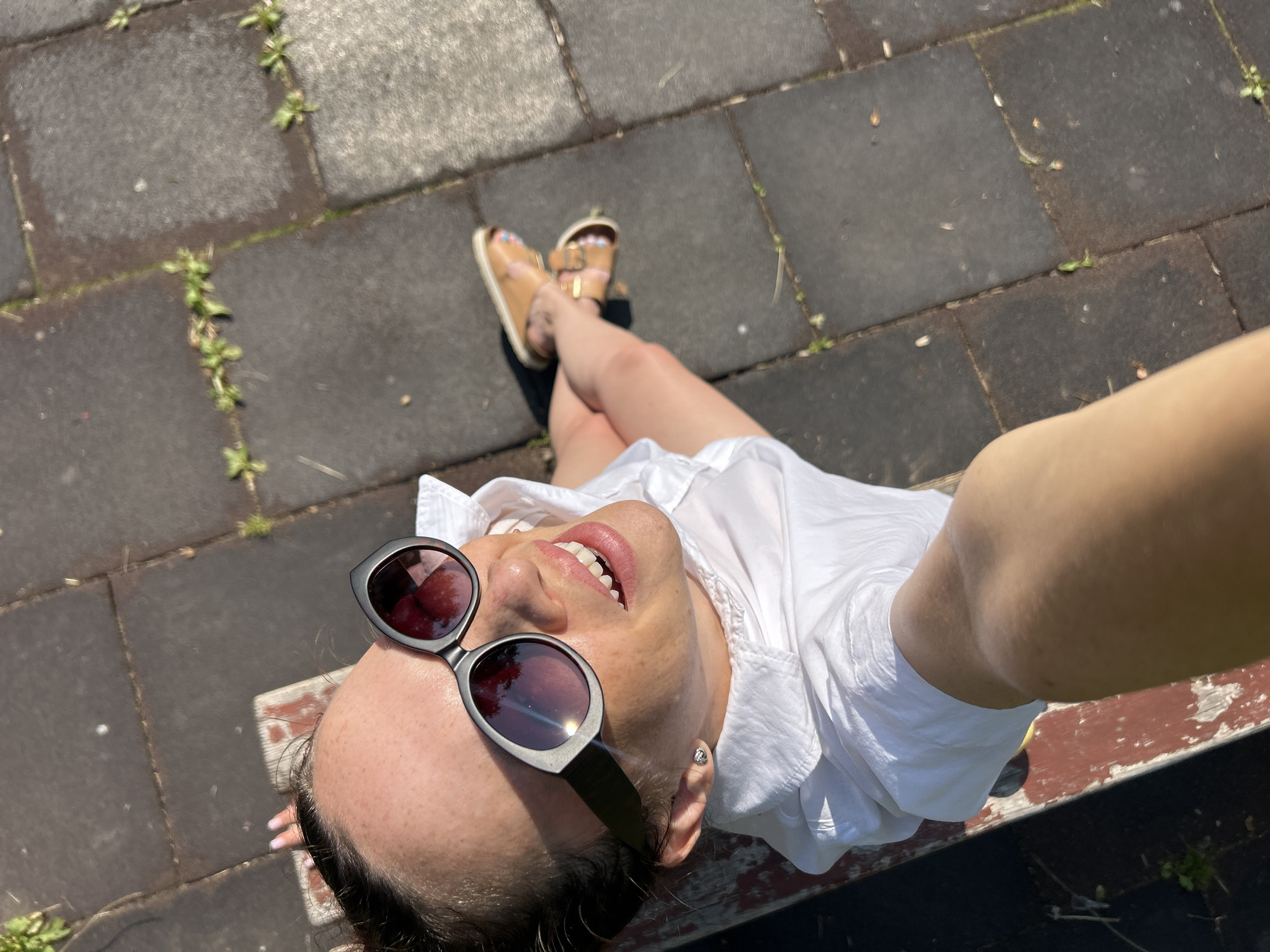 A high-angle, outdoor, medium shot captures a woman sitting on a park bench. She is looking up and to her left with a bright smile, her mouth open to reveal her teeth. She is wearing large, dark-rimmed sunglasses with reflective lenses and a white collared shirt. Her left arm is extended forward, holding the camera. Her legs are crossed, and she is wearing light brown sandals. The ground appears to be paved with dark square tiles, with some grass growing in the gaps between them. A red and white park bench is visible beneath her. The lighting suggests it is daytime, with the sun casting shadows. The location is identified as the city of Chaikovsky, Russia.