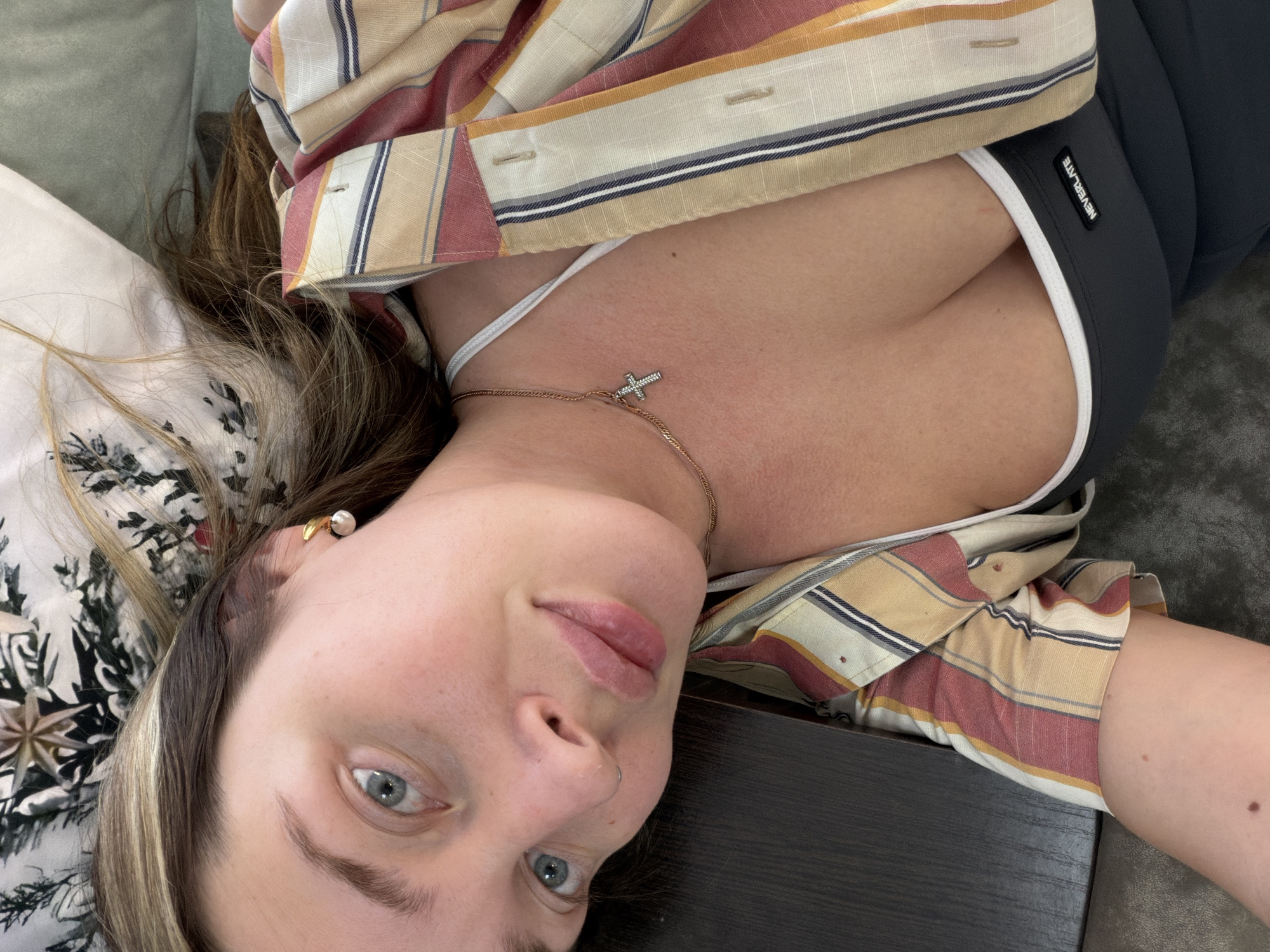A close-up, slightly tilted indoor shot features a young woman lying down, looking directly at the camera. Her fair skin, light brown hair with blonde highlights, and striking blue-green eyes are prominent. Her expression is neutral, bordering on a soft smile.
She is dressed in a multi-colored striped button-up shirt, featuring shades of red, cream, yellow, and dark blue, worn open over a white bralette or tank top. A delicate gold chain necklace with a small, sparkling silver cross pendant rests on her chest. A pearl earring with a gold-toned setting is visible in her right ear. The lower part of her visible attire includes dark gray athletic wear with white trim, on which a black tag displaying the text "NEVERLITE" is clearly seen.
Her head rests partly on a white pillow adorned with a black and white floral pattern. A dark brown or black flat surface, possibly a table, is visible to her side, and a textured gray surface, likely a couch or carpet, completes the background. The image depicts a casual, relaxed moment, possibly a selfie, within an indoor setting in Chaikovsky, Russia.