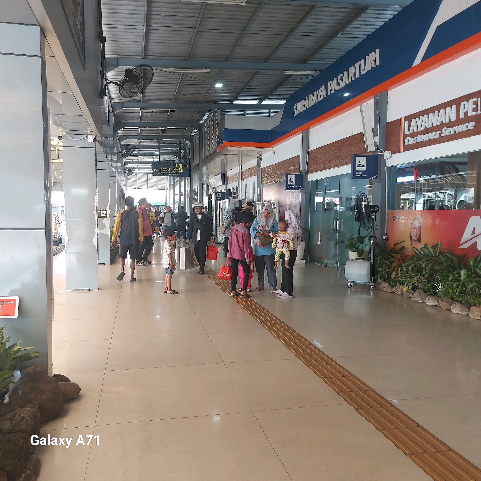 The image depicts the interior of the Surabaya Pasar Turi train station in Surabaya, Indonesia. The scene is a covered outdoor walkway or concourse of the station, with a light-colored tiled floor and a ribbed, light-colored ceiling supported by metal beams. People are seen walking and standing.
In the foreground, a young boy in a striped shirt and denim shorts stands looking to the right. To his left, there's a decorative rock formation with some greenery. Further into the scene, several individuals are visible. Two women, one in a blue hijab and another in a patterned headscarf, are holding a baby. Another woman in a black outfit with a white helmet is walking away from the camera, carrying red bags. A man in a yellow and grey t-shirt and dark shorts is walking towards the right.
Above, a prominent sign reads "SURABAYA PASARTURI". To the right of this sign, another sign indicates "LAYANAN PEL" which is likely "Layanan Pelanggan" or Customer Service, with "Customer Service" written below it in English. There are also signs for "Loket" (Ticket Counter). A standing fan with a water mister is positioned near a potted plant. Other architectural elements include pillars, glass doors, and lighting fixtures. The environment suggests a busy transportation hub. The bottom left of the image displays "Galaxy A71", indicating the device used to capture the photograph.