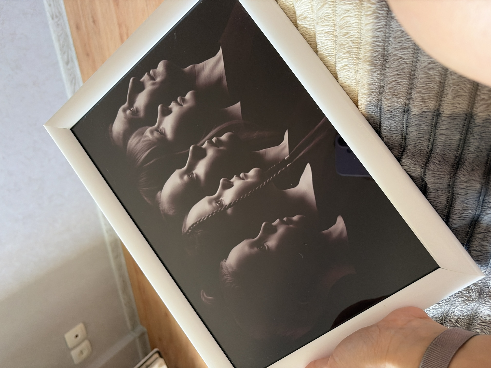The image shows a framed black and white photograph of five people in profile, stacked vertically. The frame is white. The photograph is held at an angle, with the right side slightly higher than the left. The background includes a striped textured surface and a wooden surface, likely a table or floor. The scene could be indoors, possibly in a home, but the image doesn't conclusively confirm its location in Chaikovsky, Russia, or any other city. The hand with a watch band is holding the right bottom corner of the frame.