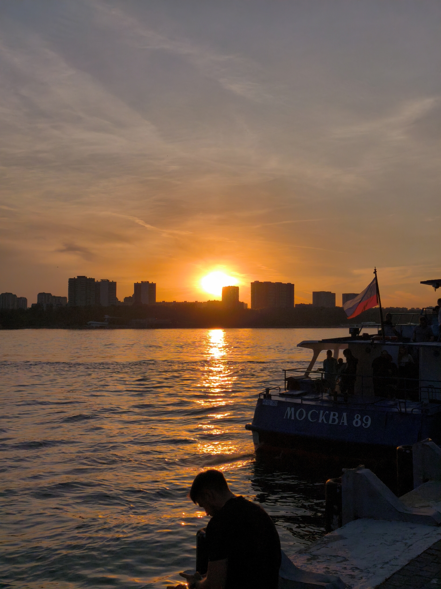This detailed image captures a vibrant sunset over a wide river, looking towards a cityscape in Moscow, Russia. The sky transitions from a soft, pale grey-blue with wispy clouds at the top to an intense, glowing orange and yellow at the horizon, dominating the central portion of the frame. The setting sun, an intensely bright, circular disc, is positioned just above the distant skyline, casting a prominent, shimmering path of golden light across the rippled river surface towards the foreground.
In the mid-ground, a line of multi-story buildings forms a dark silhouette against the brilliant sunset, interspersed with dark foliage and trees along the riverbank. A smaller, lighter-colored structure is visible at the waterline on the distant left.
In the mid-right foreground, a dark blue and white passenger boat is positioned, displaying the name "МОСКВА 89" (MOSKVA 89) in white Cyrillic letters on its hull. A Russian flag (white, blue, red horizontal stripes) flies from a pole at its stern. Several passengers are visible on the boat's upper deck, mostly as dark silhouettes.
In the immediate foreground, to the left, a lone male figure, heavily silhouetted, sits on a concrete embankment or pier. He is facing towards the right and slightly downwards, seemingly observing the water or an object in his hands. The concrete structure includes bollards and exhibits various textures and forms. The intense sunlight reflects brightly off the water, particularly along the sun's path and near the boat and foreground figure.