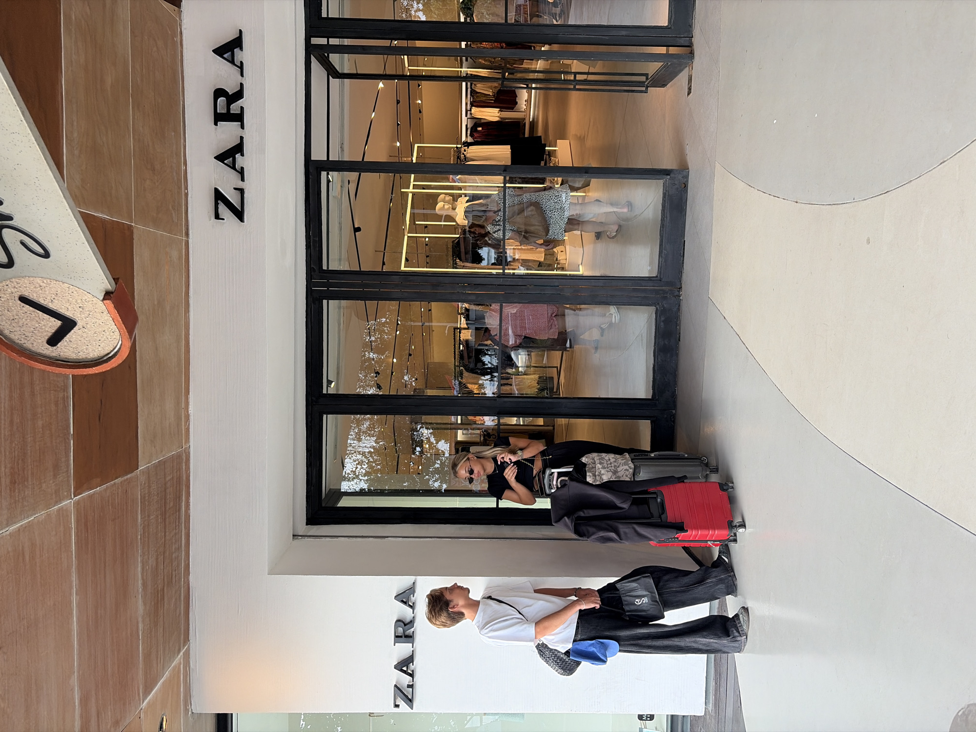 In Kuta, Indonesia, two young individuals stand outside a Zara store. On the left, a person with light brown hair, wearing a white t-shirt, black wide-leg pants, and black shoes, holds a black tote bag and a blue hat. To their right, a woman with blonde hair, wearing a black crop top and black pants, stands with sunglasses and a chain necklace. They are both looking towards each other, and several pieces of luggage, including a red suitcase and a grey suitcase, are positioned in front of them. The Zara store has large glass doors and windows, revealing clothing racks and mannequins inside. The exterior of the store features a white facade with the Zara logo prominently displayed. The ground outside the store is paved with a light-colored material, with curved lines creating a patterned walkway. A portion of the ceiling is visible, made of wooden planks.