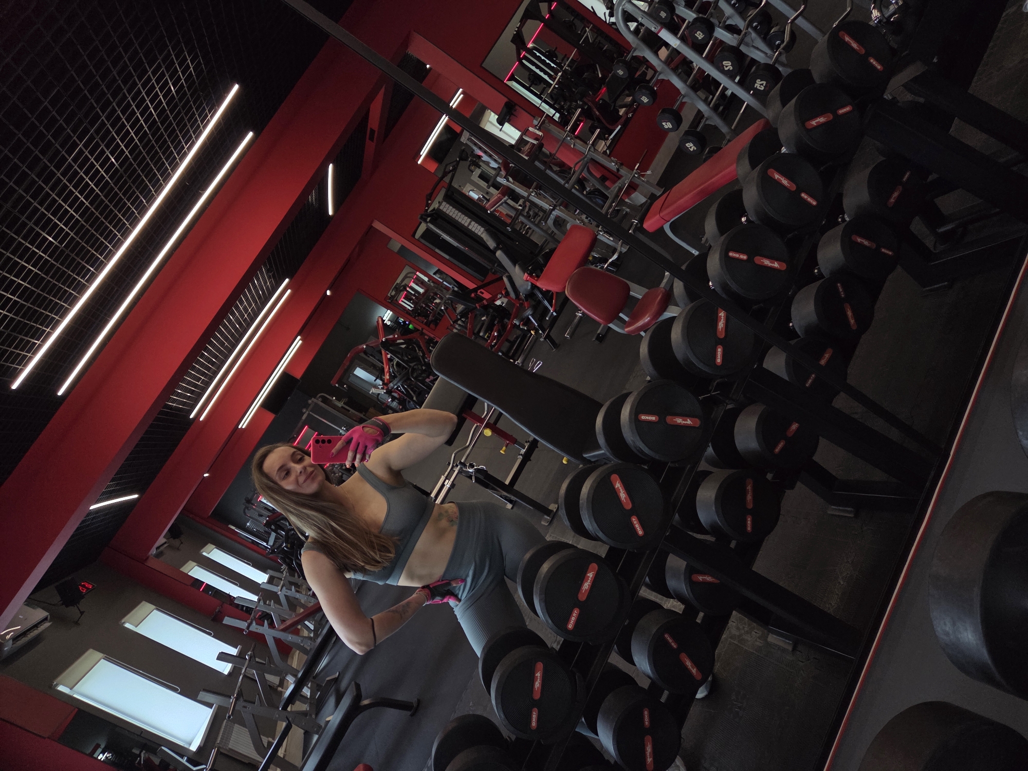 A woman with long brown hair, wearing a grey sports bra and matching leggings and pink gloves, poses for a selfie in a gym. The gym has black ceilings with white rectangular lights, red walls, weightlifting equipment, and racks of dumbbells. The image's location is in Birobidzhan, Russia.