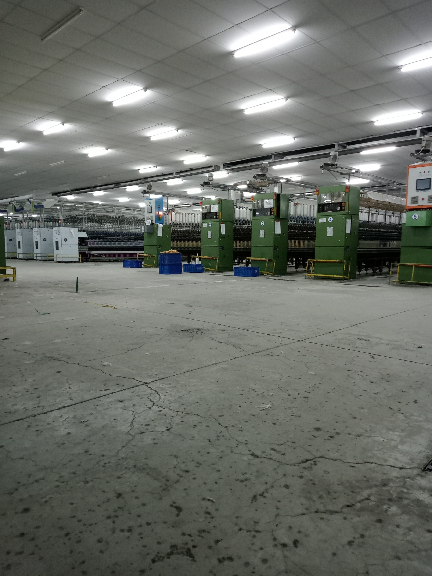 The image displays the interior of a large industrial factory floor, likely a textile or yarn production facility, located in Nglundo, Indonesia. The space is dominated by numerous rows of heavy industrial machinery, predominantly colored olive green and white, extending into the background. These machines appear to be spinning or winding units, fitted with many spools of white and off-white thread or yarn.
The floor is made of concrete, showing significant signs of wear with prominent cracks and various stains or debris scattered across its surface. Scattered intermittently between the machinery are blue plastic crates or baskets, and yellow metal safety barriers or frames are positioned in front of some equipment. Numbers "0", "1", "2", "3", "4", and "5" are clearly visible on the front panels of the green machines, likely serving as identification markers. Control panels are also visible, including a blue unit with an orange indicator light on the left and an orange-faced panel with a digital screen on the right.
The ceiling consists of a grid of light-colored panels, brightly illuminated by numerous long, rectangular fluorescent light fixtures, which are the primary source of light in the facility. There are no people visible in the image, suggesting the machinery is currently idle or the factory is not in active operation. The absence of people and the bright artificial lighting mean the time of day and external weather are indeterminate. The overall scene conveys a functional, albeit currently quiet, industrial environment.