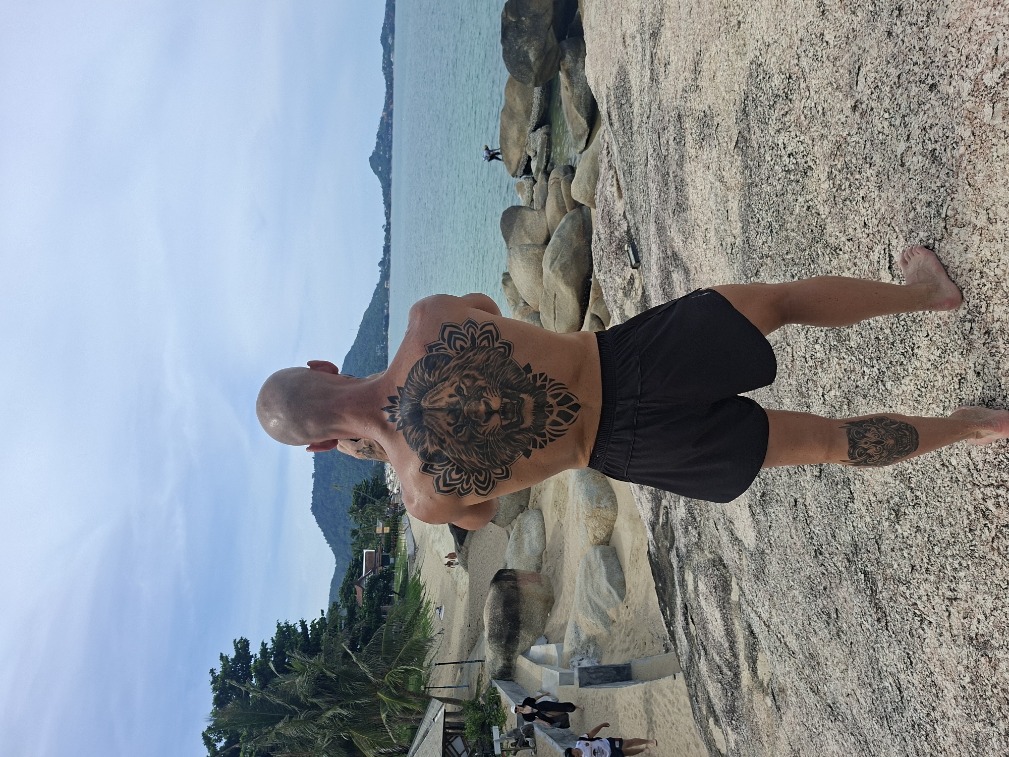 A man stands on a rocky shore, his back to the camera, facing a tranquil blue ocean. He has a large, intricate lion tattoo covering his back, and a smaller tattoo of a dragon head on his left calf. The man wears black shorts and is barefoot. In the background, palm trees line a sandy beach, with buildings and lush green hills rising in the distance. The sky is partly cloudy, with a light haze. Other people are visible on the beach and in the water, appearing small due to the distance. The location is Baan Hua Thanon, Thailand.