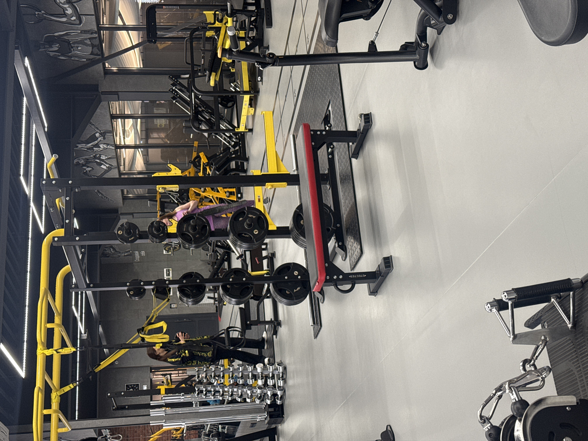 The image shows the interior of a gym, likely in Votkinsk, Russia, given the Cyrillic text reflected in a mirror. There is gym equipment including racks with weights, dumbbells, and various workout machines. Two individuals are present, one lying on a bench under a weight rack and another near a set of dumbbells. The gym's color scheme incorporates black and yellow. The lighting includes overhead lights and linear LED strips, contributing to a bright and modern aesthetic. The walls are painted a neutral color, possibly gray or white.