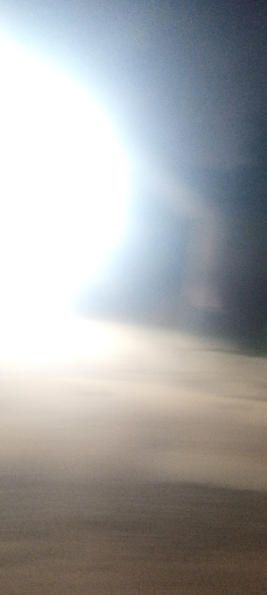 The image appears to be a landscape view with a prominent light source, likely the sun, heavily overexposed on the left side. The right side of the image is darker, with a gradient transitioning from bright to dark. A band of lighter, possibly hazy or cloudy, material stretches across the mid-ground of the image, separating the bright upper area from a darker lower area. The image lacks specific details to identify the location as Narayi, Nigeria, or any other discernible features indicative of a specific geographic location or human activity. The absence of identifiable objects or people indicates an impersonal and featureless scene.