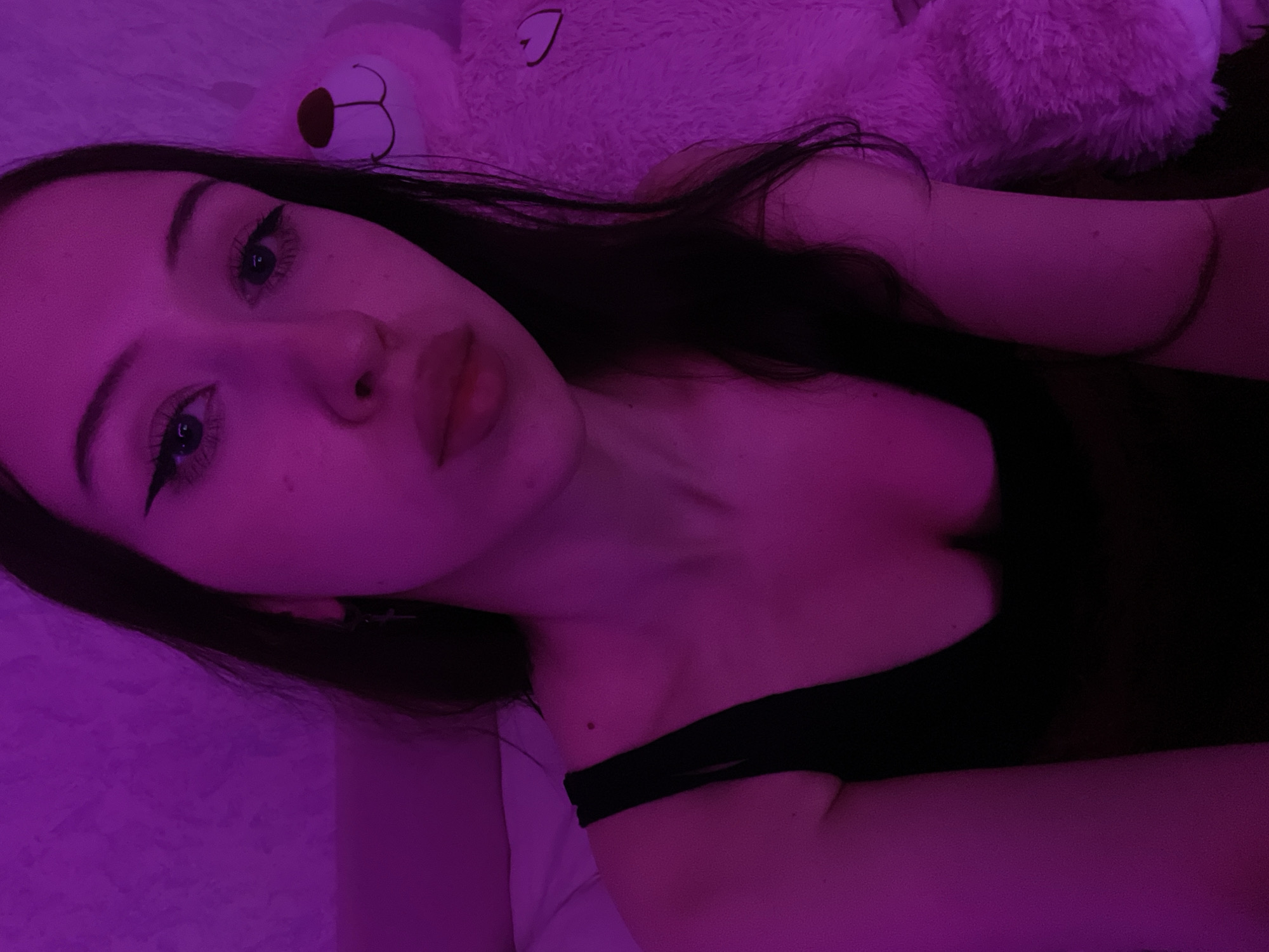 A young woman with long, dark hair and prominent winged eyeliner is depicted in a close-up, selfie-style shot. She is wearing a black tank top. The scene is illuminated by a vibrant pink light, casting a strong hue over her skin and the surrounding environment. To her right, a large, fluffy, pink teddy bear is partially visible. The background appears to be a textured wall, also bathed in the pink light. The setting suggests an indoor environment, likely a bedroom. There is no discernible connection to the city of Votkinsk, Russia, in the provided image.
