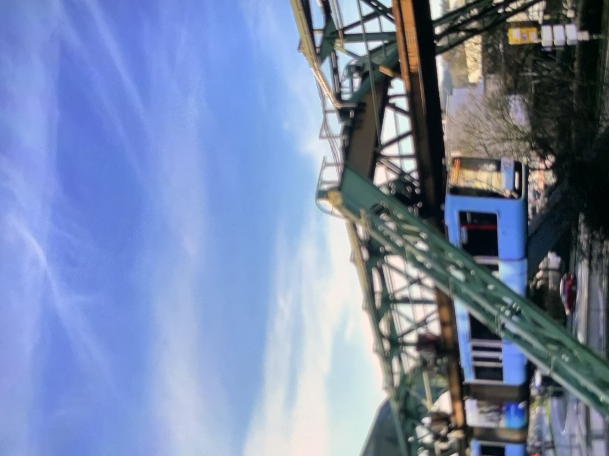 A blue monorail train travels on an elevated track against a bright blue sky with wispy white clouds. The track structure is green and metallic, with visible supports and beams. Below the track, there are trees and some buildings. A red car is visible on a road in the lower part of the image. The scene appears to be in an urban environment.