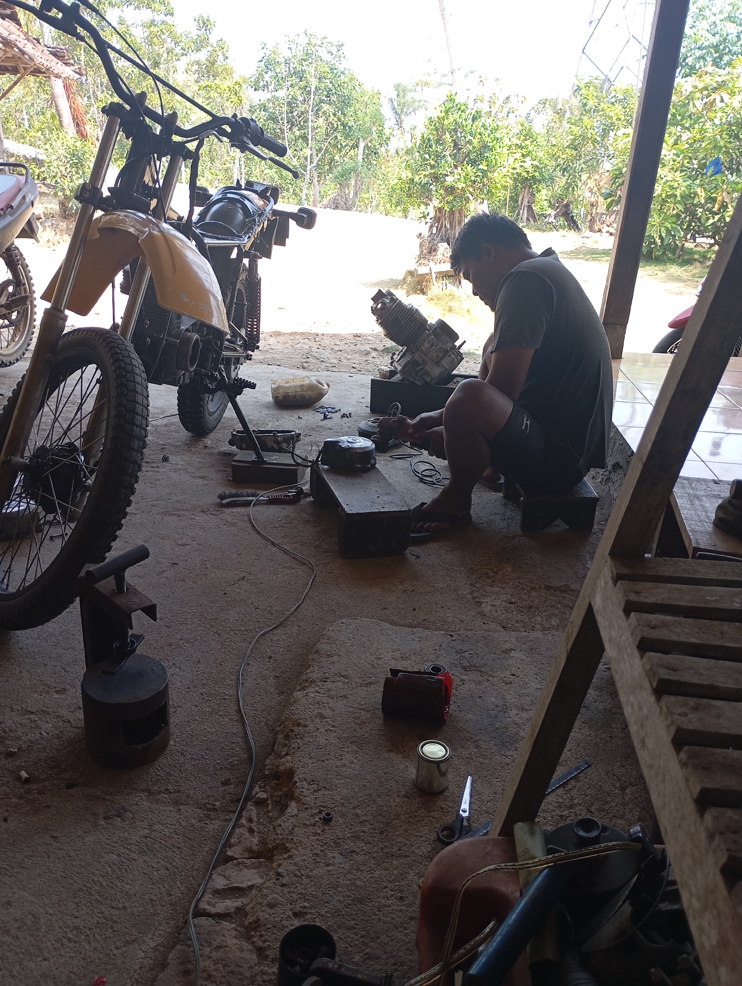 A man is working on a motorcycle engine in a workshop located in Ketepung, Indonesia. The man, wearing a dark t-shirt and shorts, is seated on a small wooden stool and appears to be focused on the engine parts in front of him. Several motorcycle parts are scattered around the workshop, including a yellow front fender and a wheel visible in the foreground. Tools and other miscellaneous items, such as a can of paint, scissors, and electrical wires, are also present on the concrete floor. The workshop appears to be an open-air structure with a wooden roof and pillars. Lush green trees and vegetation are visible in the background, suggesting a rural or semi-rural setting. The overall scene depicts a typical motorcycle repair or maintenance activity in an outdoor workshop.