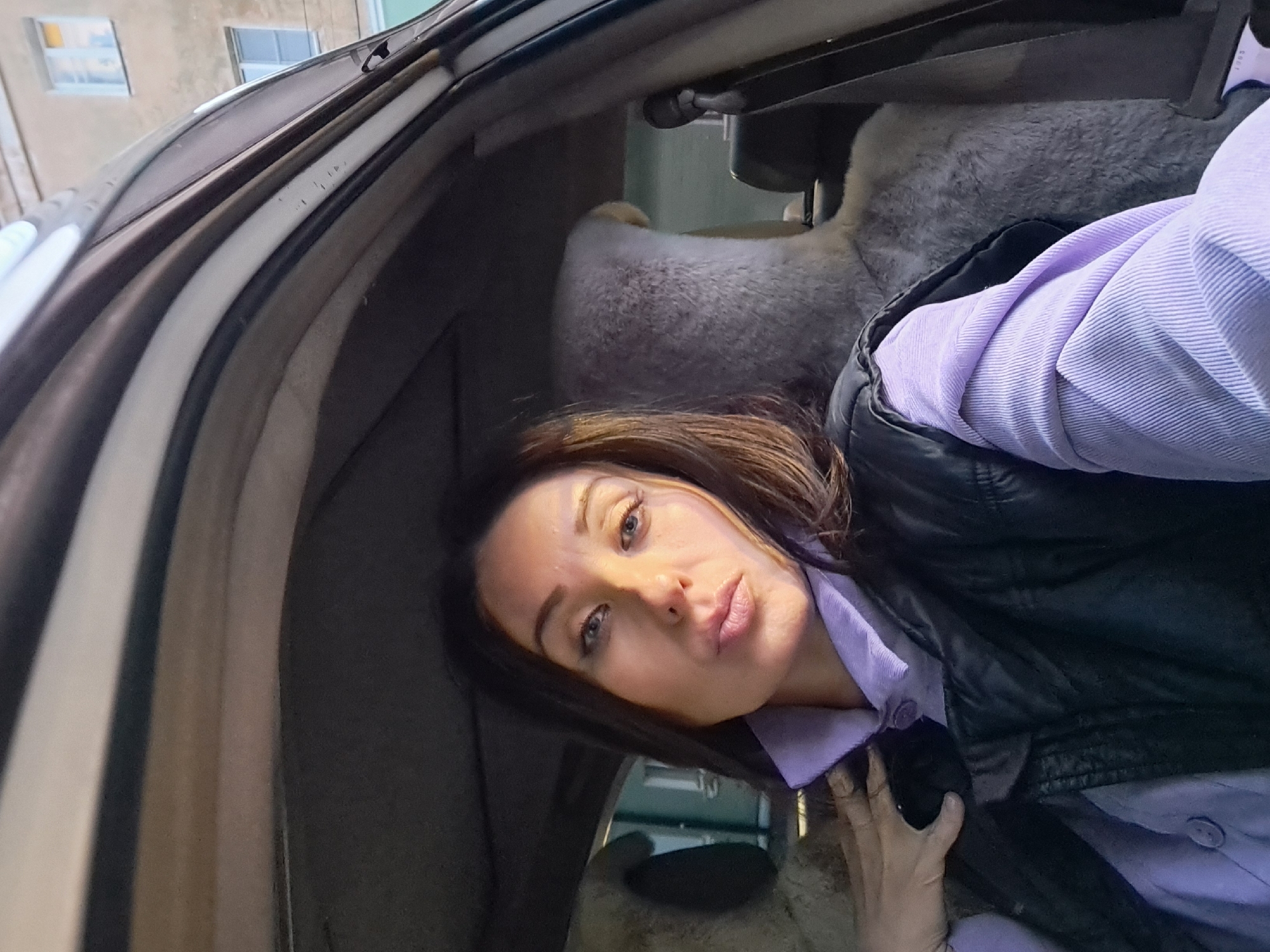 A woman with long, dark hair, wearing a lavender collared shirt and a black vest, is taking a selfie inside a car. Her lips are pursed, and her right hand is resting on her left shoulder. The car's interior is visible, including a fluffy gray seat cover in the background. Outside the car, a building with windows is visible, suggesting an urban setting. This scene is located in Irkutsk, Russia.