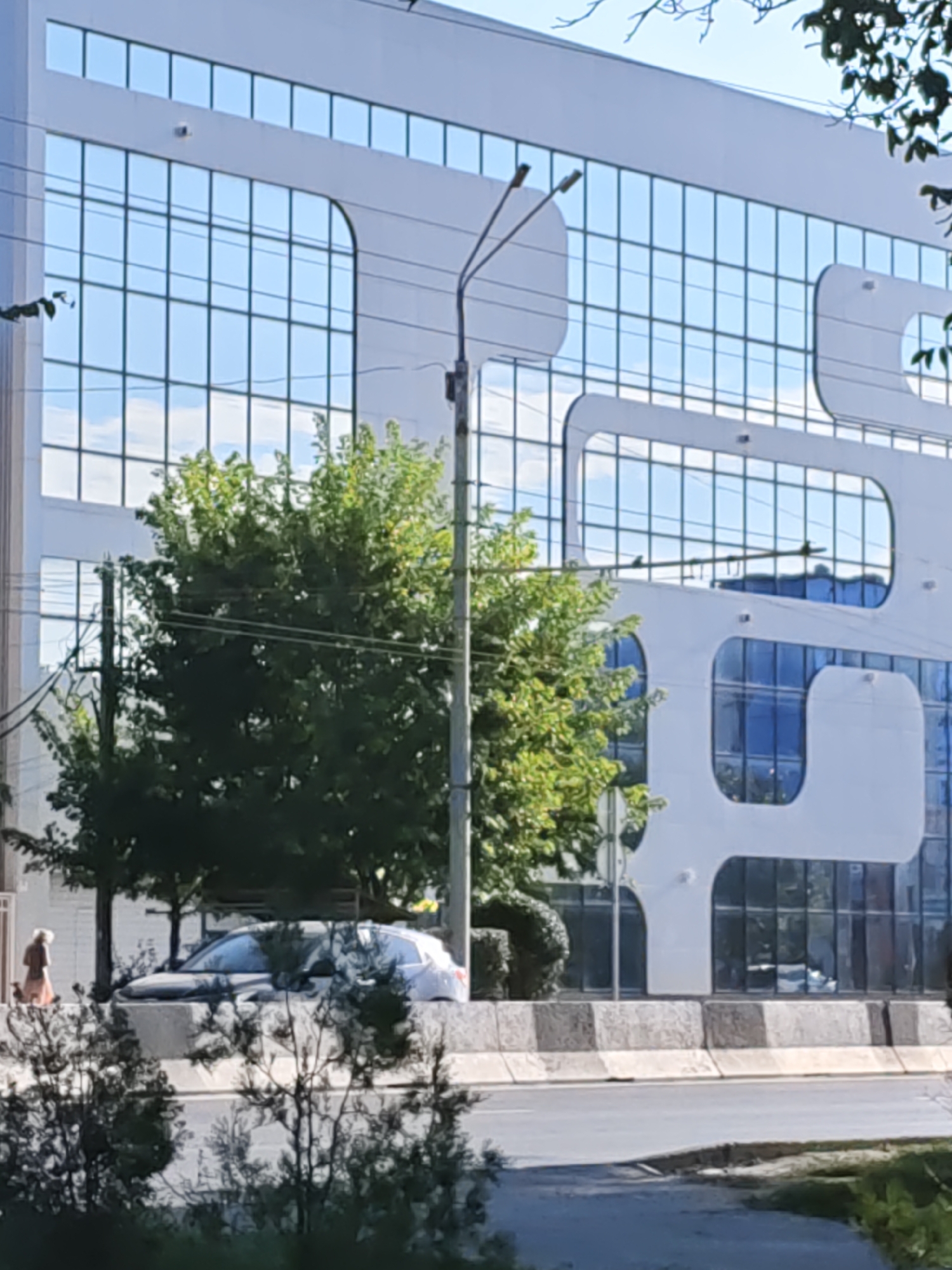 The image captures a modern building with a distinctive white facade and large, reflective glass windows in Rostov-on-Don, Russia. The building features a unique design with curved white elements integrated into its structure. In front of the building, there are trees and a street with a car visible. A person is walking on the sidewalk next to the street. The scene is set during the daytime, with clear sky visible through the building's windows.
