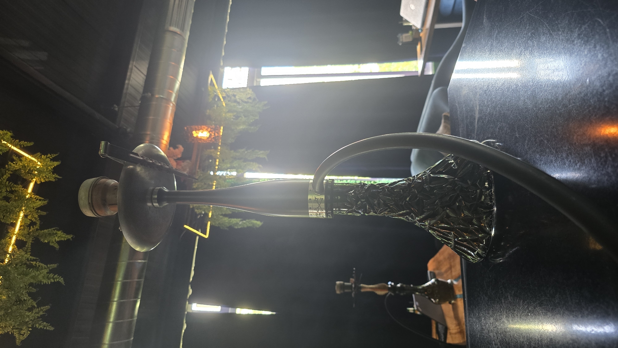 The image shows a hookah in an outdoor setting. The hookah is metallic with a dark, textured base and a black hose. There are decorative lights and trees visible in the background, suggesting an outdoor bar or lounge. In the background, there are buildings, and signs suggesting this could be a commercial area. Another hookah is visible in the background as well. A person is partially seen in the background. Without additional information or landmarks, pinpointing the exact location in Sofia, Bulgaria, is not possible based on the visual information alone.