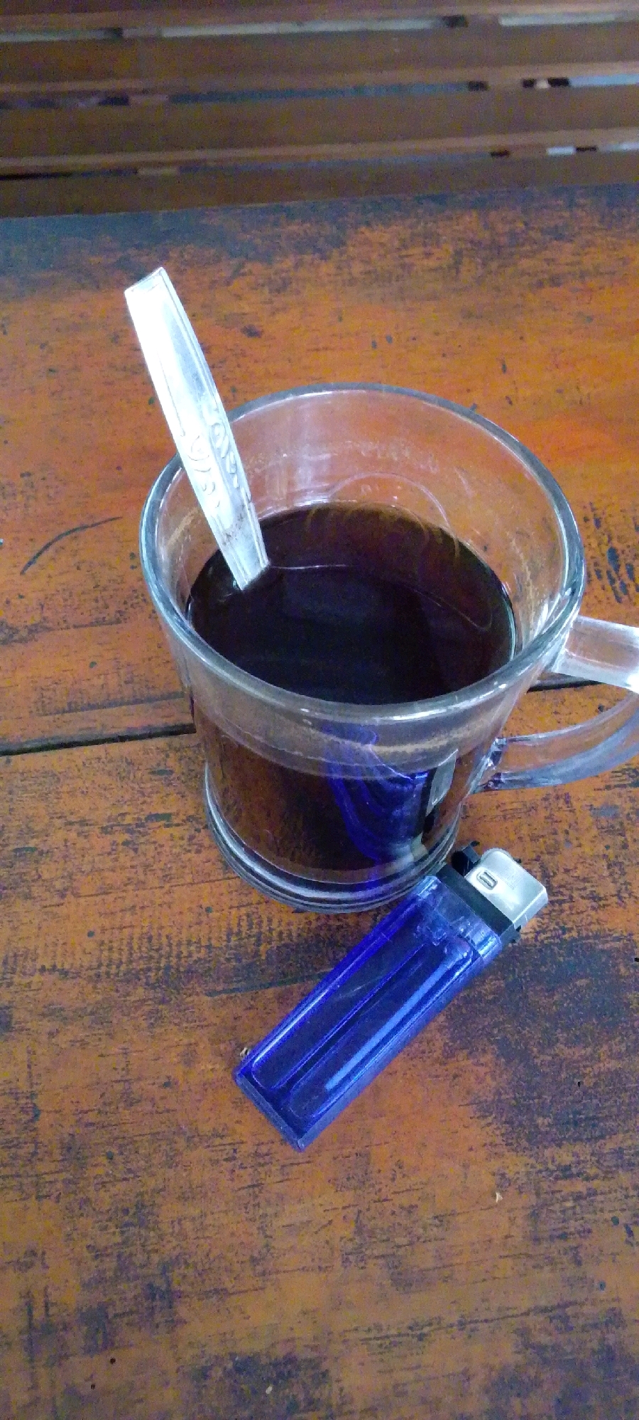 An overhead, close-up shot shows a clear glass mug filled with dark coffee, a silver spoon resting inside, and a blue lighter on its side, all placed on a rustic wooden table. The table surface has a weathered, grainy texture with visible wood grain and some dark speckles. The mug is positioned on the right side of the frame, with the spoon angled towards the top left. The blue lighter lies horizontally in the lower right quadrant of the image. The background above the table is out of focus, revealing horizontal wooden planks. The scene appears to be in an outdoor or casual setting, possibly a cafe or home.