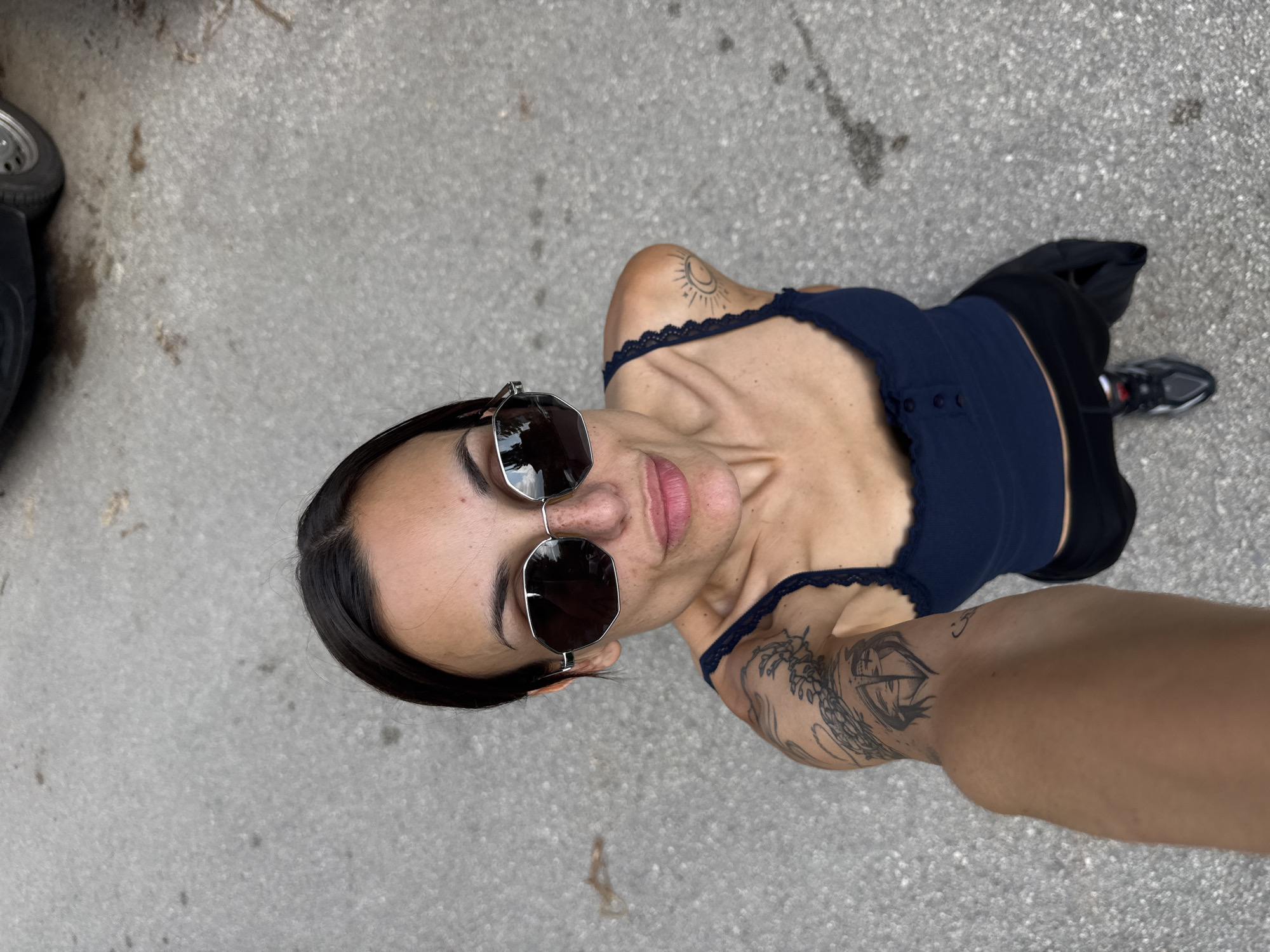 A woman is featured in a high-angle selfie photograph, taken outdoors in Pleven, Bulgaria. She has dark hair, parted in the middle and pulled back, and wears silver-framed, octagonal sunglasses with dark lenses that reflect the sky and display "Polaroid" on the left lens. Her lips are slightly pursed, and freckles are visible on her nose and cheeks. She wears a dark blue camisole with lace trim along the neckline and three small buttons on the front. Tattoos are visible on both arms: a sun and crescent moon design on her right shoulder and a larger, intricate blackwork tattoo depicting abstract figures within a shield-like outline on her left upper arm. Her right arm is extended downwards and slightly left, holding the camera. She wears dark trousers and a dark athletic shoe with red and white accents on her left foot. The background consists of light grey asphalt or concrete pavement, marked with scattered dark stains and minor organic debris. A dark vehicle, specifically a tire with a silver hubcap and part of a fender, is visible in the upper right background.