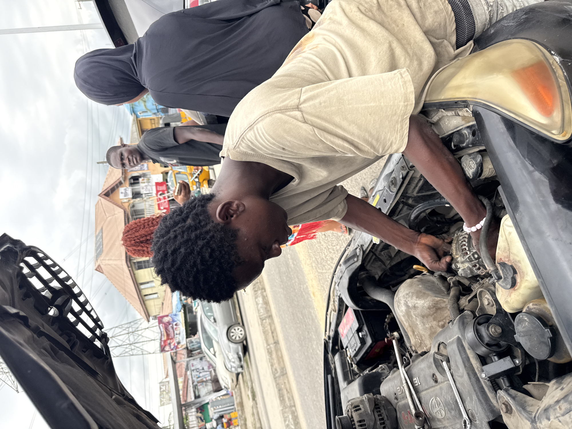 A medium shot captures a young man in Ado, Nigeria, deeply engrossed in repairing the engine of a car. He is positioned over the engine bay, his hands working meticulously on the internal components. The car's hood is open, revealing the complex arrangement of the engine. Two other individuals stand nearby, observing the repair process. The scene is set on a city street, with buildings and other vehicles visible in the background. Overhead power lines add to the urban context.