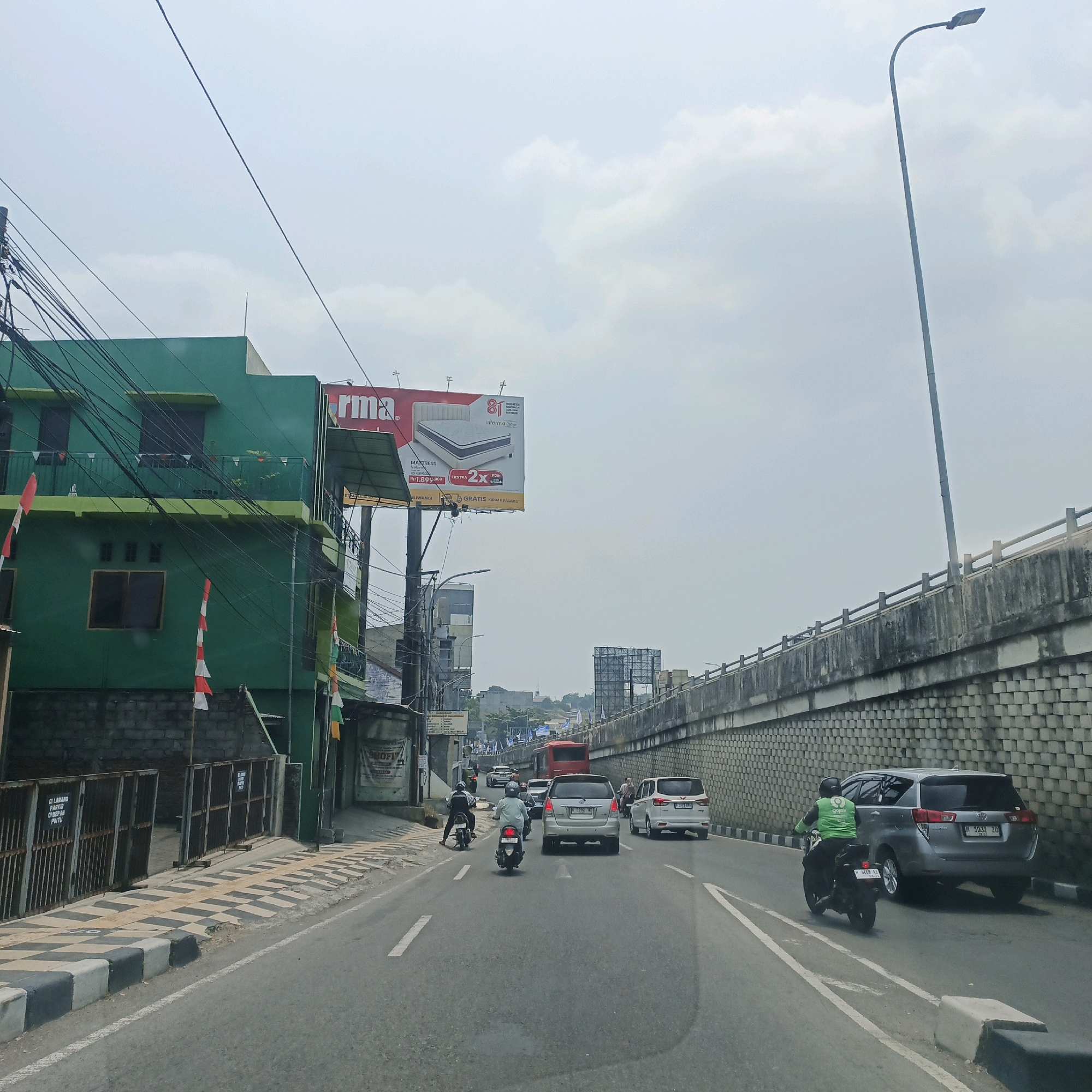 The image depicts a street scene in the city of Semarang, Indonesia, likely on a cloudy day. The perspective is from a vehicle traveling on a multi-lane road. On the left, a green building with multiple floors is visible, adorned with Indonesian flags. Above the building, a large billboard advertising a mattress is mounted. The street itself is paved and marked with white lines. Several vehicles, including cars and motorcycles, are present on the road, with some traffic. A person on a motorcycle wearing a green jacket is visible in the foreground on the right. To the right of the road, a concrete barrier with a textured pattern runs along an elevated road or bridge structure. A tall street lamp is also prominent in the sky. The overall impression is of a typical urban environment with active traffic.