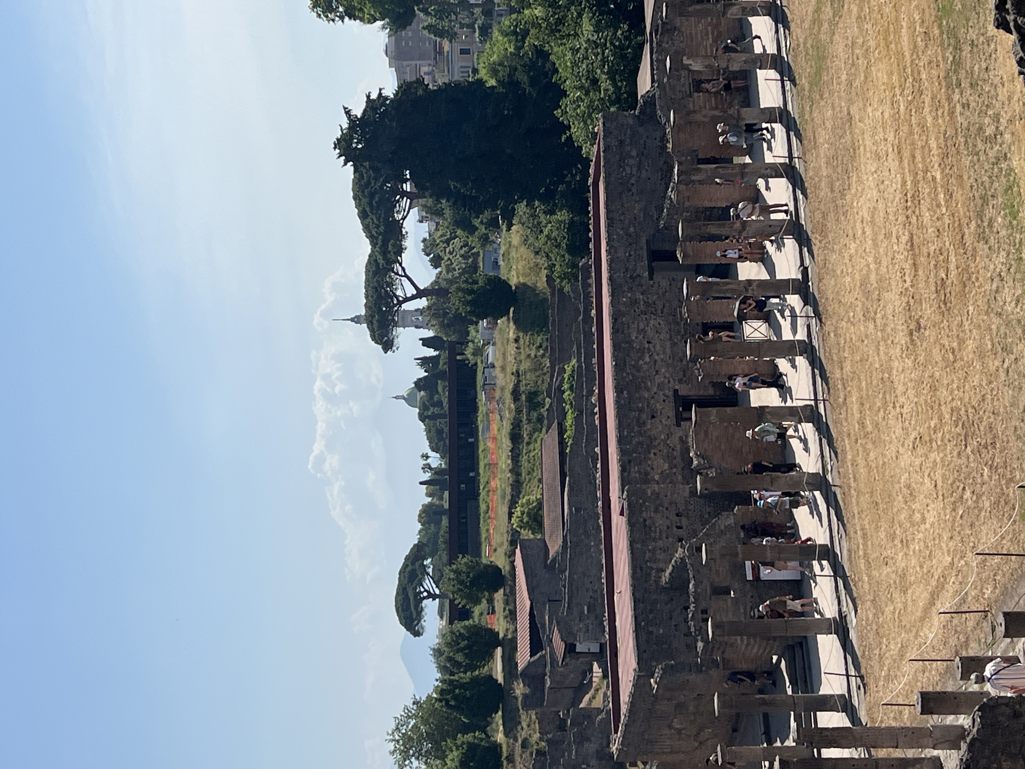 The image captures a view of the ancient Roman ruins of Pompeii, Italy, under a clear blue sky with scattered clouds. In the foreground, weathered stone structures with red-tiled roofs are visible, suggesting well-preserved ancient buildings. A colonnade of tall, cylindrical stone pillars lines a walkway, casting distinct shadows on the ground. Several tourists, dressed in casual summer attire, are scattered throughout the scene, some walking along the colonnade, others observing the ruins. The ground in front of the colonnade is covered in dry, golden grass or straw, suggesting an archaeological site undergoing maintenance or preservation.
In the mid-ground, more ancient stone buildings are visible, some with their roofs intact and others partially ruined. Orange construction fencing is present in some areas, indicating ongoing work. The landscape behind the ruins is dotted with lush green trees, including distinctive umbrella-shaped pine trees.
In the background, a prominent green dome is visible, likely belonging to a church or historical building. Further in the distance, a tall, slender tower with a pointed spire rises above the treeline, possibly a bell tower or monument. The backdrop features rolling hills and mountains, with a hazy atmosphere suggesting distance. The overall impression is one of historical significance, with modern visitors exploring the remnants of an ancient civilization.