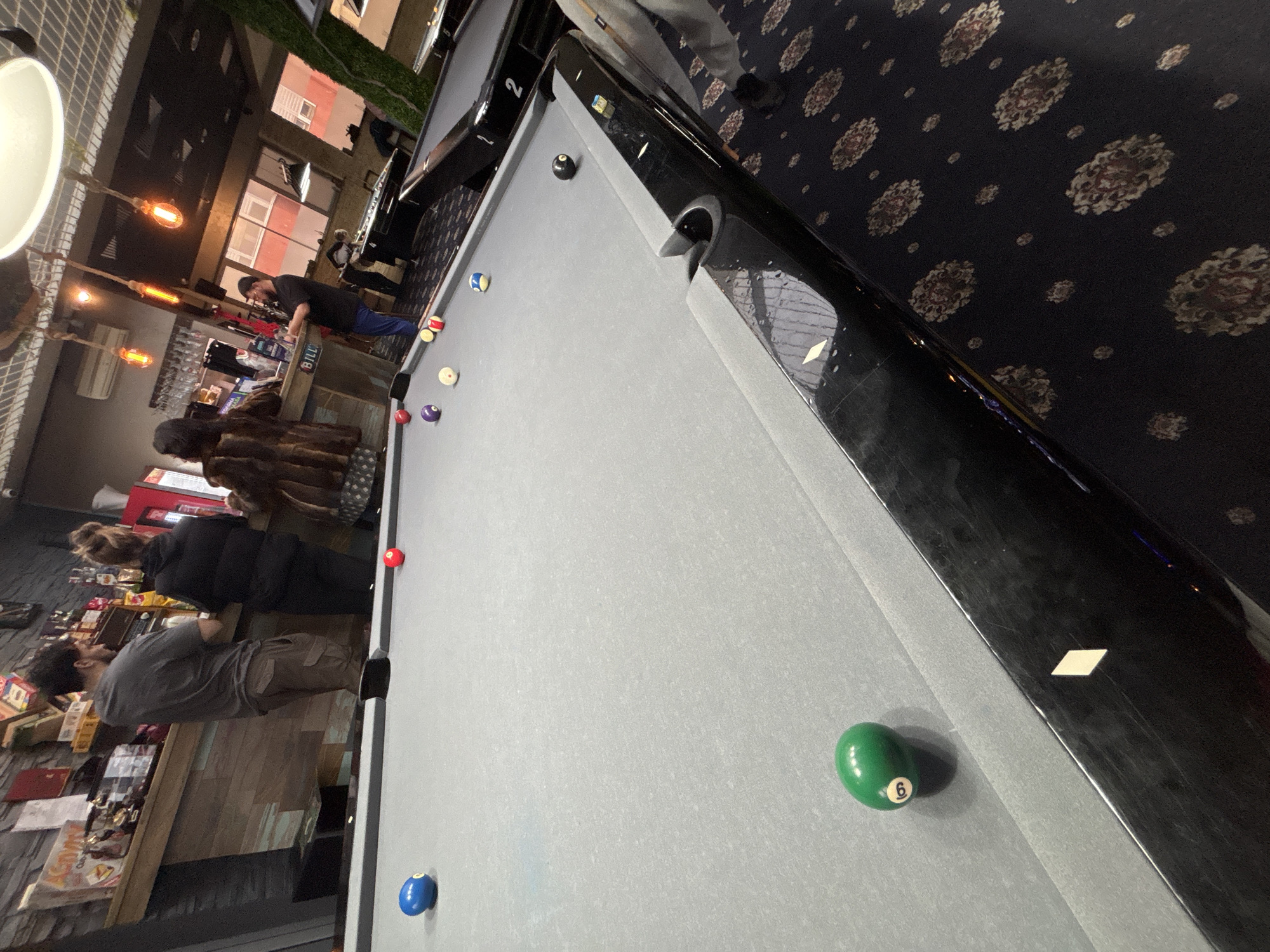 The image depicts the interior of a bar or recreation area, likely located in Plovdiv, Bulgaria, with multiple people standing around a pool table. The pool table is in the foreground, partially obscuring the rest of the scene. Several pool balls are scattered on the table's grey surface, including a green ball with the number 6 visible. People are standing at the side of the pool table, watching or preparing to play. The background reveals a bar area with a counter, shelves, and more people. Lighting fixtures hang from the ceiling. The room has a relaxed and social atmosphere. A carpet with a patterned design is also visible in the background. Another pool table is visible towards the top left of the frame, with the number "2" displayed on it.