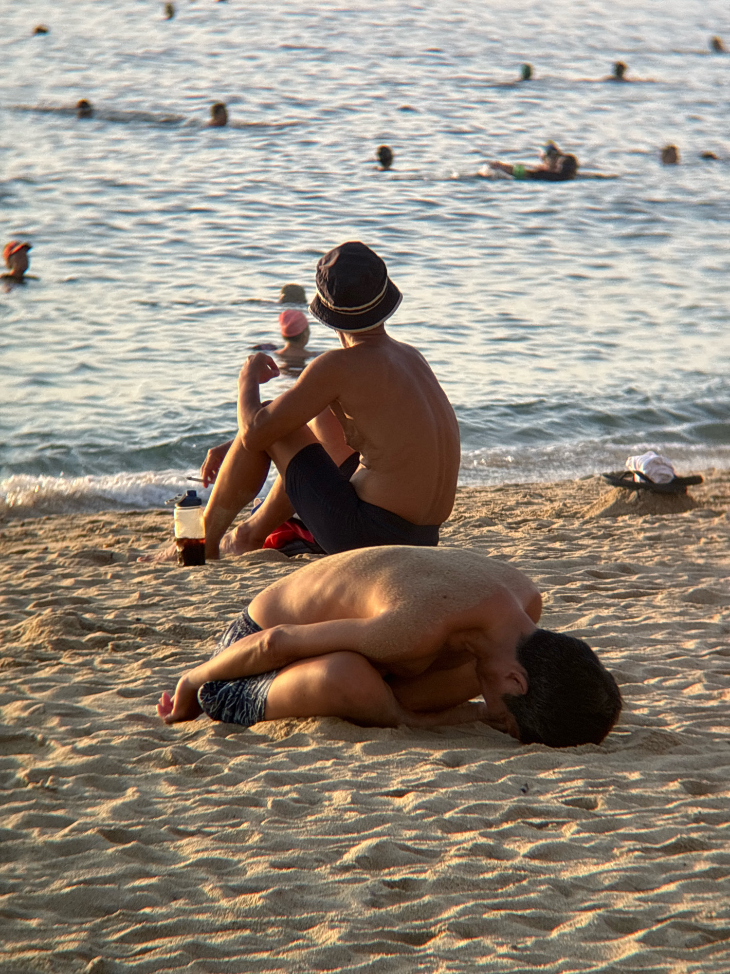 Two men are on a sandy beach with the ocean in the background. The man in the foreground is lying on his side, facing away from the camera, with his knees drawn up. The man in the background is sitting up, facing away from the camera, with his legs crossed. There are other people in the water in the background. The sun is setting, casting a warm glow on the scene. This is likely Nha Trang, Vietnam.