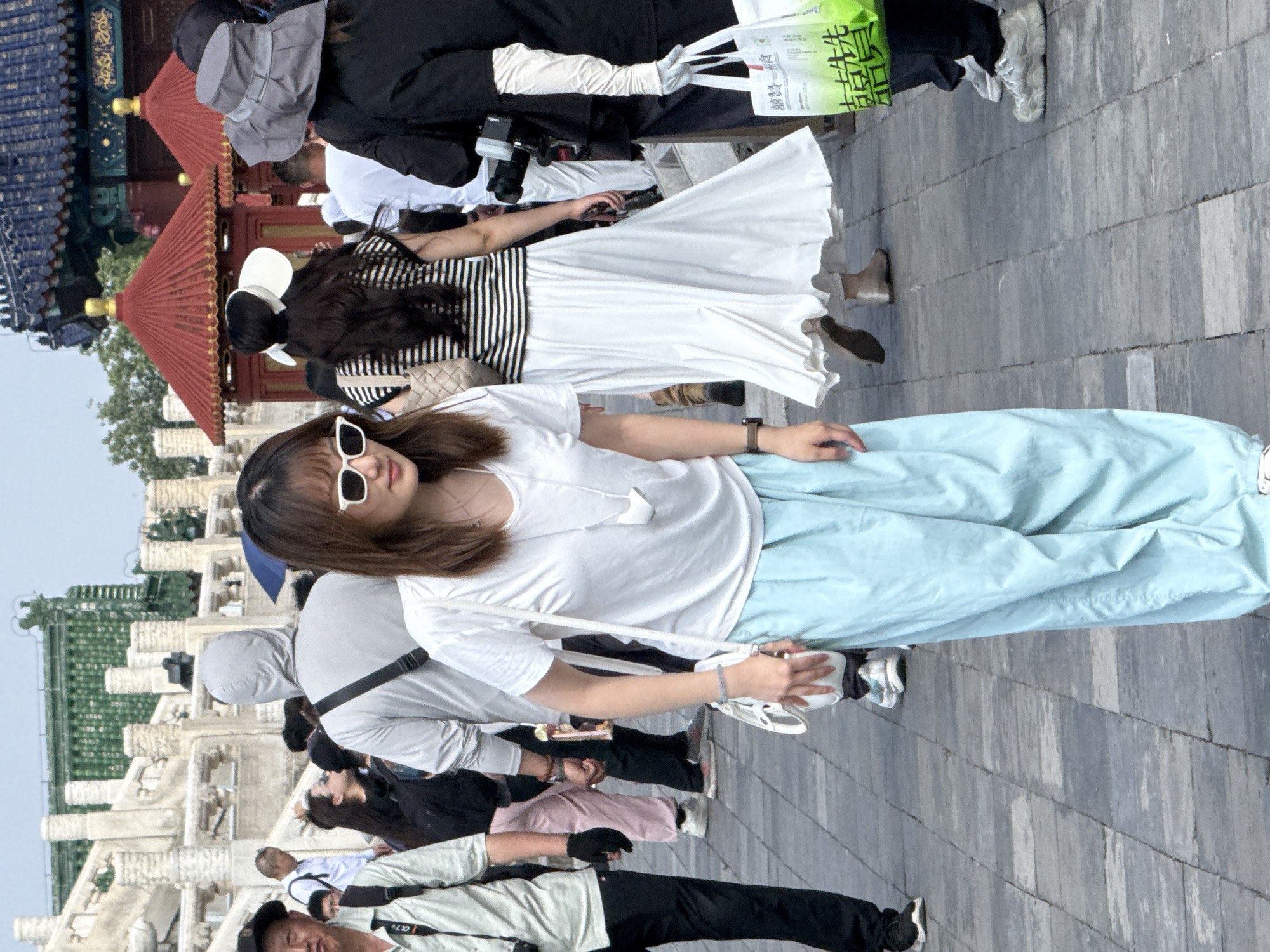 A medium shot captures a crowd of people walking on a paved pathway in front of traditional Chinese architecture, likely in Dongcheng District, China. The foreground is dominated by a young woman wearing a white t-shirt, pale blue loose-fitting pants, and white-framed sunglasses. She carries a white purse on her left shoulder and has a bracelet on her left wrist. Behind her, other individuals are visible, including a woman in a black and white striped shirt and a white skirt, and a person in a black jacket and a wide-brimmed grey hat holding a white plastic bag with green Chinese characters. The architecture in the background features ornate eaves and red pavilions. The overall scene depicts a public space with tourists or visitors exploring a culturally significant area.