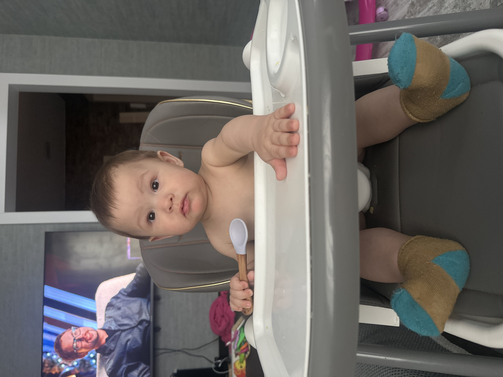 A baby wearing brown and blue striped socks sits in a high chair, holding a wooden spoon. The baby's face is directed towards the camera with a neutral expression. In the background, a television screen displays a man wearing glasses and a blue shirt. The scene appears to be indoors, with the high chair positioned in front of a grey wall and a doorway.