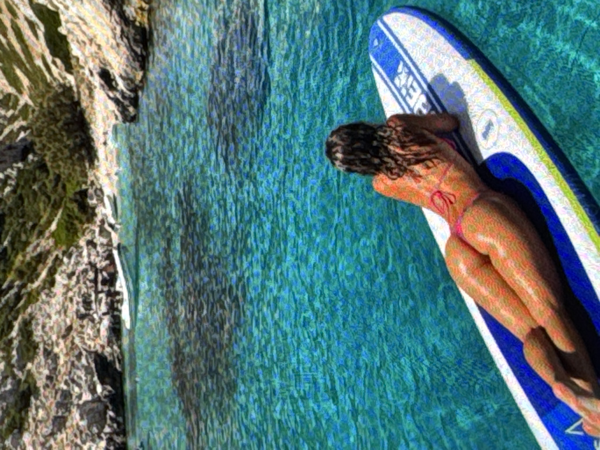 A woman lies prone on a paddleboard in clear blue water, her back to the viewer. She is wearing a pink bikini. The paddleboard is white with blue accents and lettering. The water is calm and shows the rocky shoreline in the distance, dotted with greenery. A white boat is also visible in the background, anchored near the shore. The scene appears to be a tranquil day at the beach or a secluded cove.