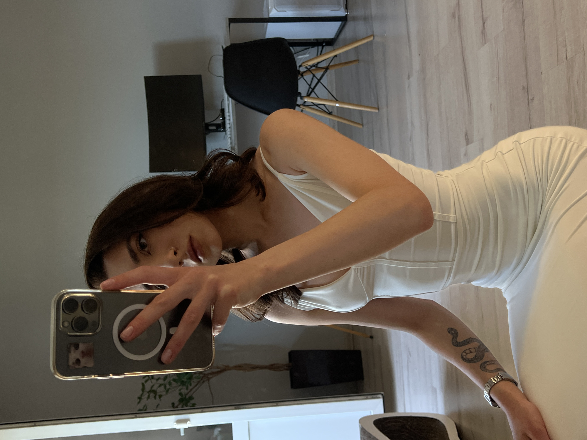 A young woman with dark hair is taking a selfie in a room with wooden floors. She is wearing a white, form-fitting dress with spaghetti straps. On her left forearm, there is a tattoo of a snake, and she is wearing a silver watch. The room appears to be a living space or bedroom, with a computer monitor and a black chair visible in the background, as well as a tall potted plant to her left. The lighting suggests it is daytime, and the overall atmosphere is casual and personal. The provided crops focus on specific elements like the phone, the woman's face, her arm tattoo, and parts of the background.