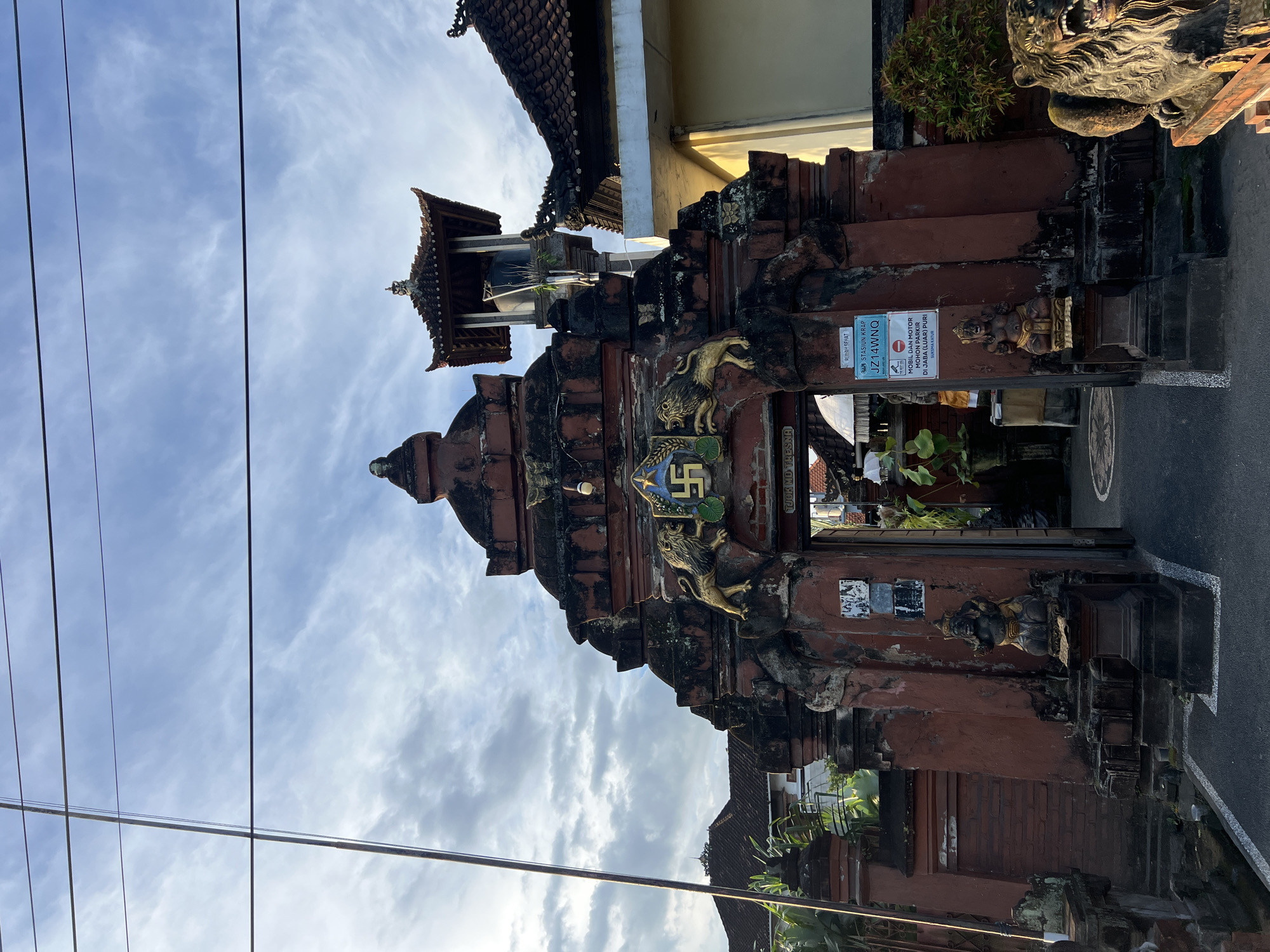 The image shows a weathered, ornate gate structure in Singapadu, Indonesia. The gate is primarily red with dark accents, constructed of brick and stone. It is adorned with carved figures, including lions and a symbol resembling a swastika. Above the gate is a small tower-like structure with a tiled roof. Several signs are attached to the gate, including one with the text "TJOK MD TRESNA." The surrounding environment includes greenery, a glimpse of nearby buildings, and power lines against a cloudy sky. The overall scene conveys a sense of cultural heritage.