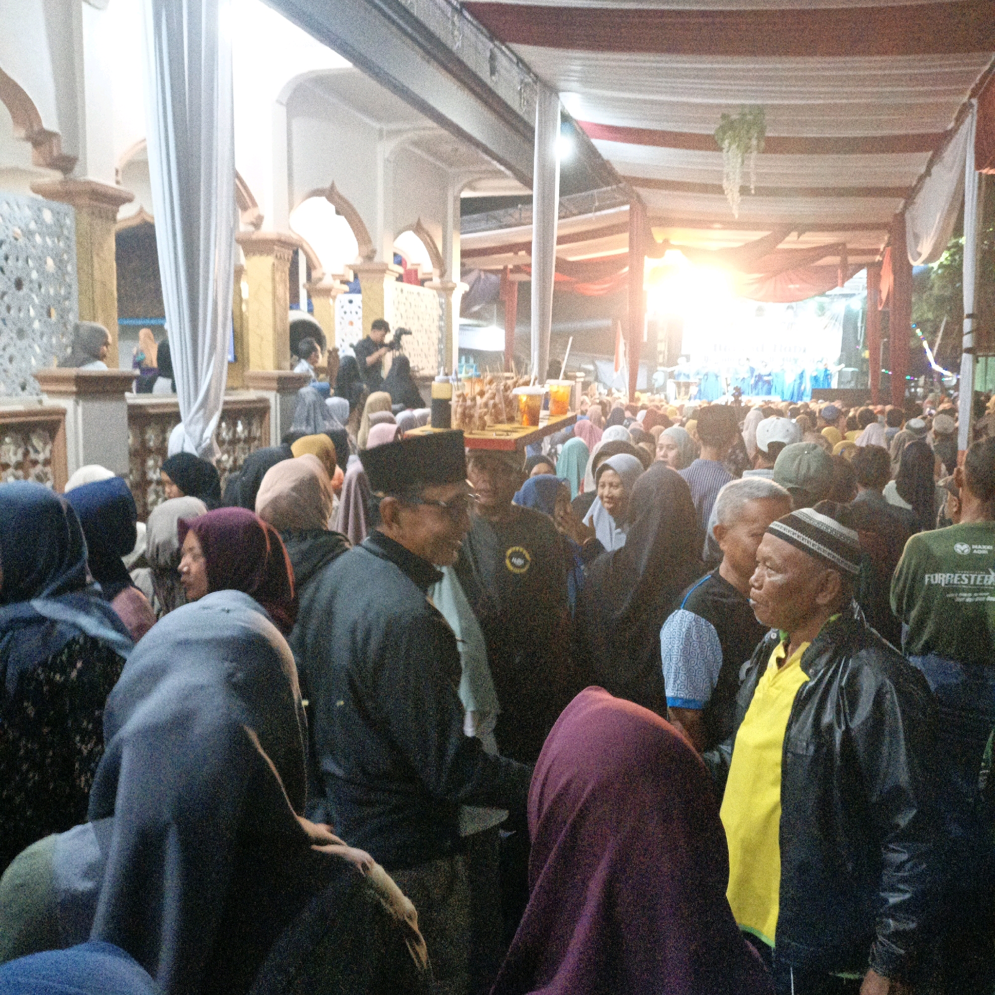 This media file captures an outdoor gathering, possibly a religious or cultural event, in Kalitorong, Indonesia. A large crowd of people, predominantly dressed in modest attire including hijabs and caps, are seen in the foreground and midground. The scene is illuminated by bright, warm light, suggesting it is late afternoon or early evening, and the weather appears to be clear.
The setting features architectural elements with arches and decorative pillars, possibly part of a mosque or a traditional building. Elaborate fabric draping, in white and red, is suspended overhead, creating a festive atmosphere. In the midground, a stall with food items and drinks is visible, indicating a communal meal or refreshment area. Further back, a stage or performance area is illuminated by spotlights, with a group of people visible on it, suggesting an entertainment or religious ceremony is taking place.
The mood seems lively and communal, with people congregating and interacting. Some individuals are looking towards the stage, while others are engaged in conversation. There is no visible text that can be clearly discerned in the image. The overall impression is one of a vibrant community event.