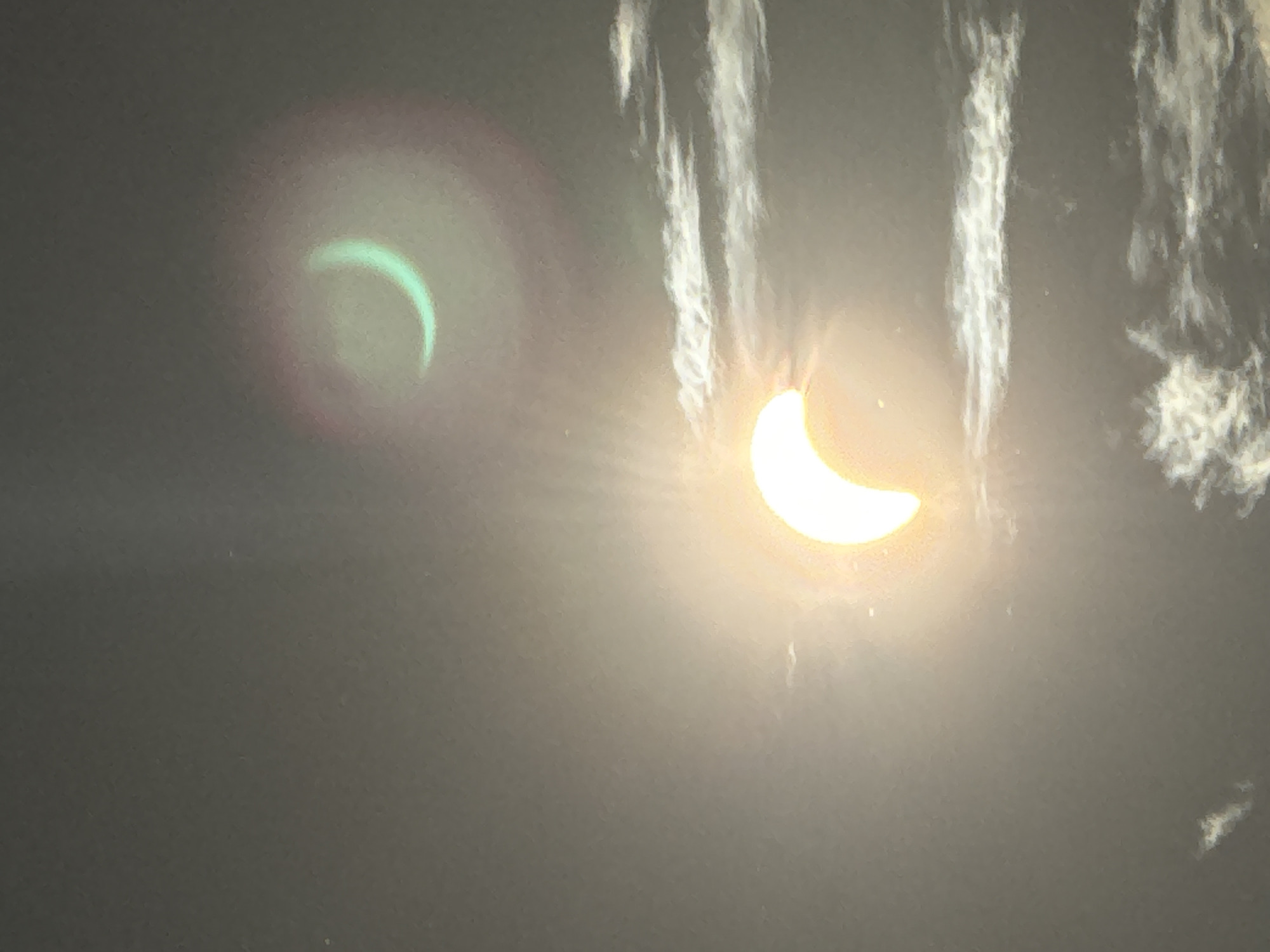 The image shows a cloudy sky with two crescent shapes. The lower crescent is large, bright yellow, and appears to be the sun partially obscured. Wispy clouds are visible below and around it. The upper crescent is smaller, has a greenish hue, and is surrounded by a faint pinkish-red halo. The overall scene is atmospheric and suggests a celestial event, possibly an eclipse, occurring in Waterford, Ireland.