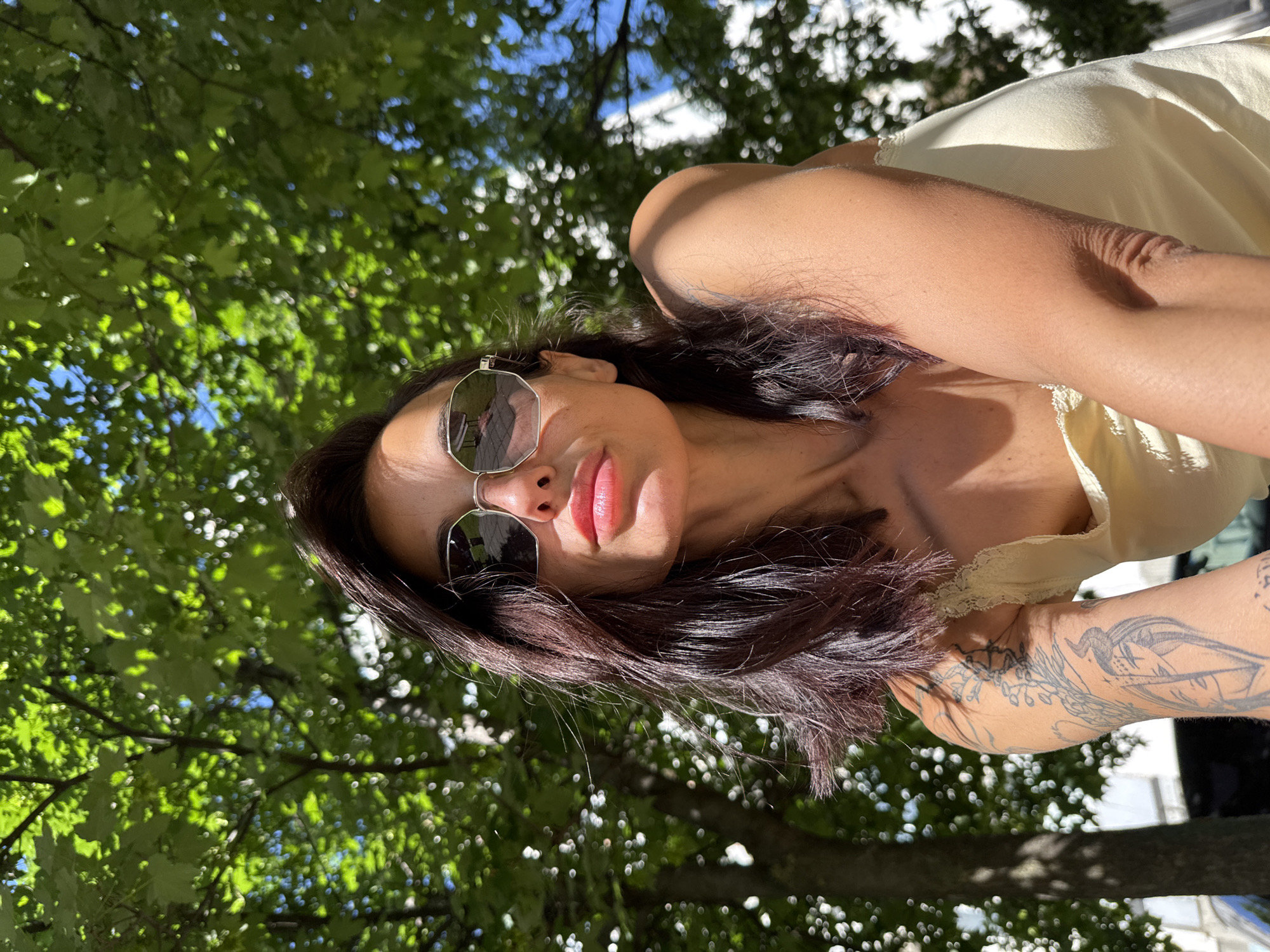 A woman with sunglasses is outdoors, possibly in the city of Pleven, Bulgaria, based on the provided context. She is wearing a light-colored top with lace detail. Her hair is dark, and she has tattoos on her arm. Green trees are visible in the background. The photo is taken from a low angle, looking up at the woman.