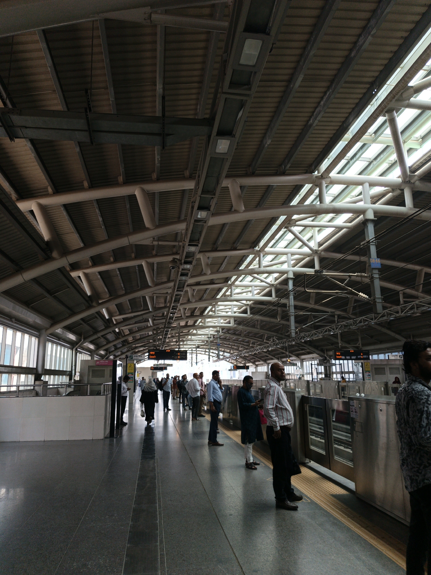 The image depicts the platform of a metro rail station in Dhaka, Bangladesh. People are standing and waiting on the gray, shiny platform. In the background, a display screen shows the coach number. The station's roof is high, with a complex network of metal beams and pipes. Skylights allow natural light to filter in. Turnstiles are positioned along the platform's edge.