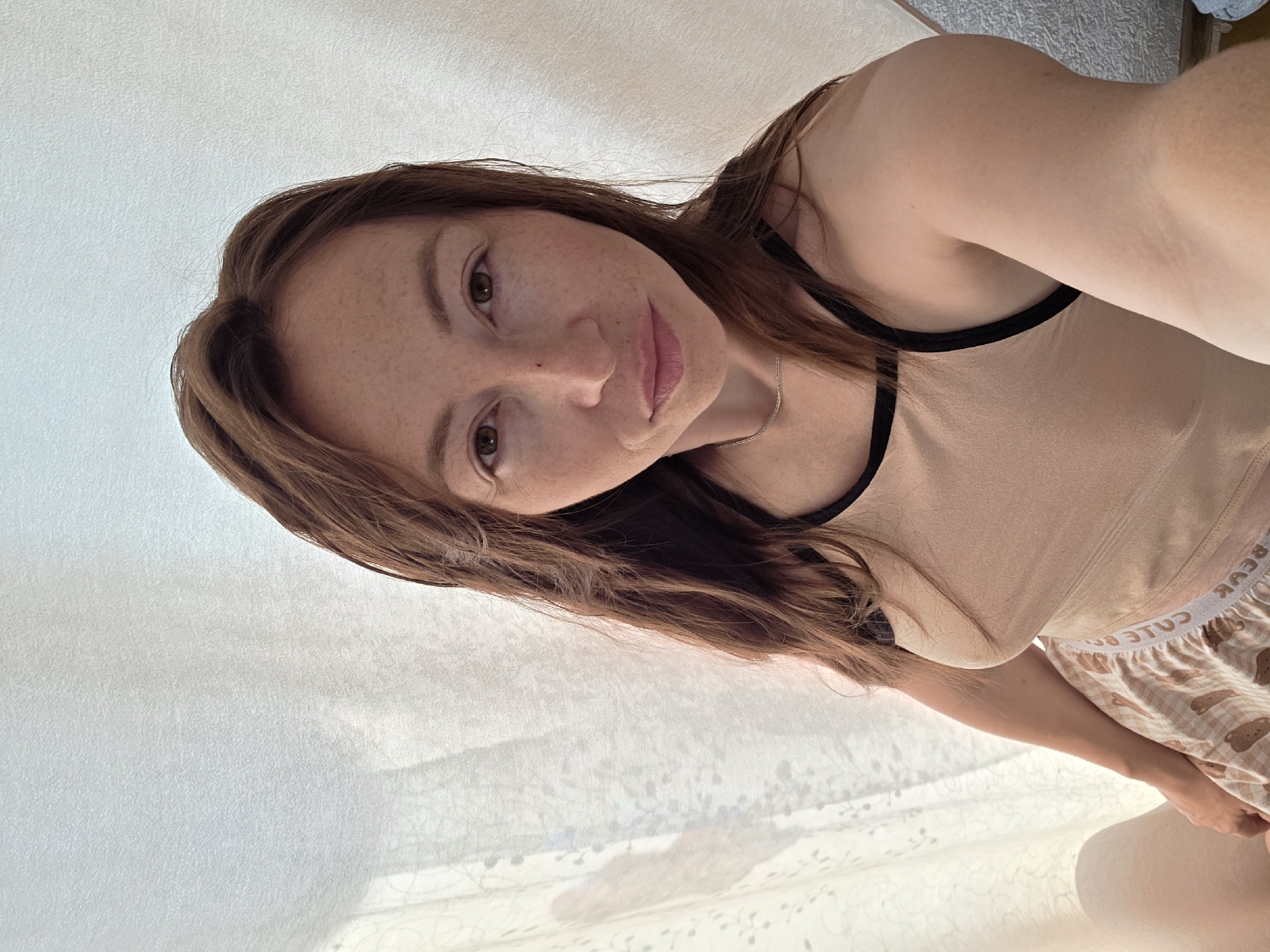 A woman with long brown hair and freckles is looking at the camera. She is wearing a light brown tank top with black trim and checkered shorts. She is sitting down with her arm resting on her leg. The background is a white curtain with a pattern. There is a shadow of a hand on the curtain to the left of the woman. The lighting is soft and natural.