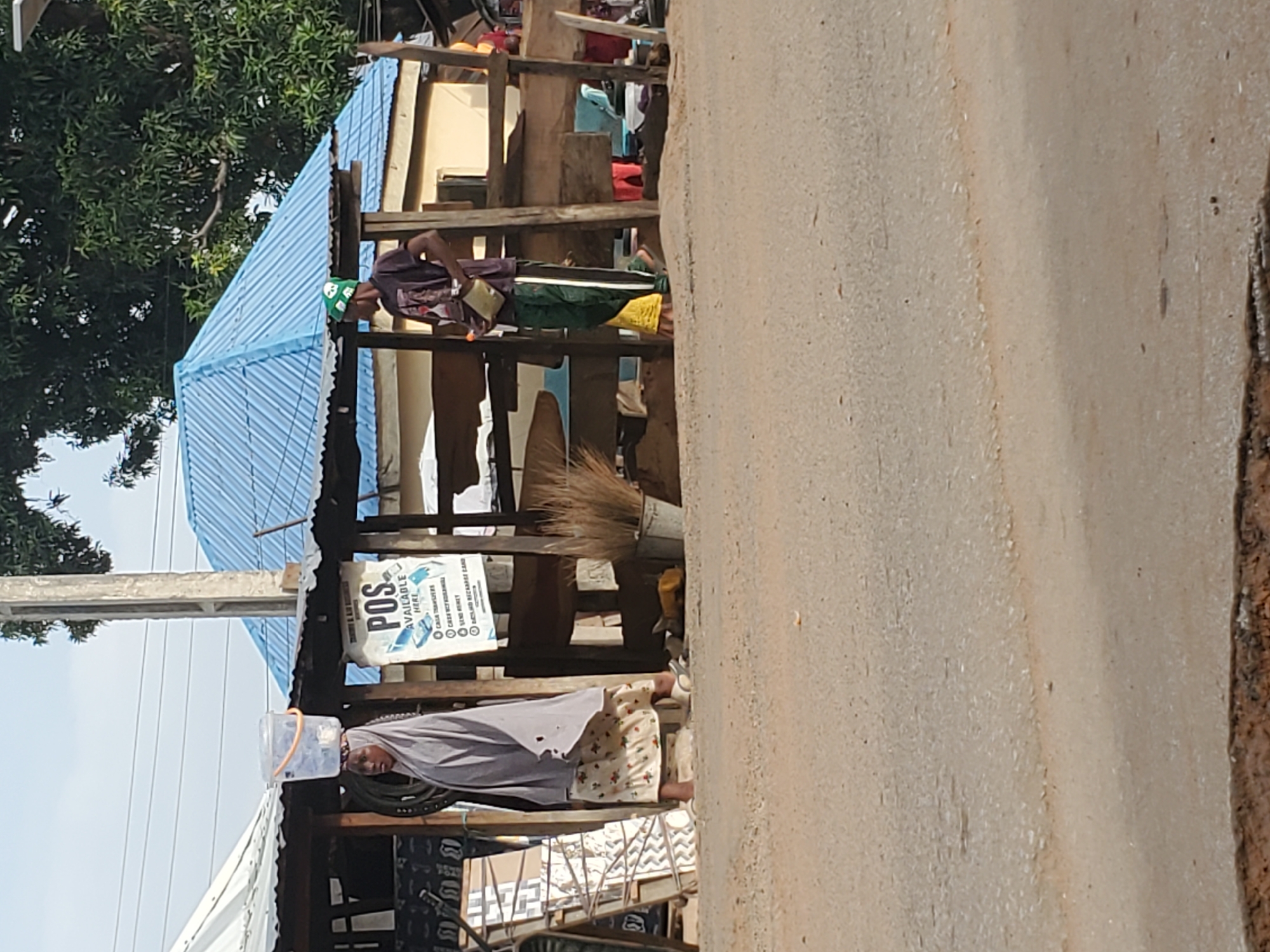 This image captures a bustling roadside or market scene in Mararaba, Nigeria, under bright daylight. The scene is dominated by makeshift commercial structures constructed from wooden frames and featuring corrugated metal roofs, some painted blue and others appearing grey or white. Lush green trees are visible in the background on the left, suggesting a verdant environment.
In the foreground on the left, a woman wearing a light-colored head covering (possibly a hijab or headscarf) and a patterned skirt stands near one of the stalls. She carries a clear plastic container balanced on her head, a common method of transport in the region. Directly to her right, a white sign with blue text is prominently displayed, advertising "POS AVAILABLE HERE" and listing services including "CASH WITHDRAWAL," "CASH TRANSFERS," "FUND ACCOUNT," "E-MONEY," and "AIRTIME RECHARGE CARD," indicating a financial service point within the market.
Further into the market, beneath a vibrant blue corrugated roof, another individual, possibly a vendor, is visible attending to a stall. This person is wearing dark clothing and a green cap with a white logo or design. Various goods and items are scattered on and around their stall. Leaning against one of the wooden supports is a traditional broom made of natural fibers.
The right side of the image shows a segment of a light-colored dirt or paved road. The clear sky in the upper left suggests fair weather. The overall impression is that of a typical, lively outdoor market or commercial area in a Nigerian town.