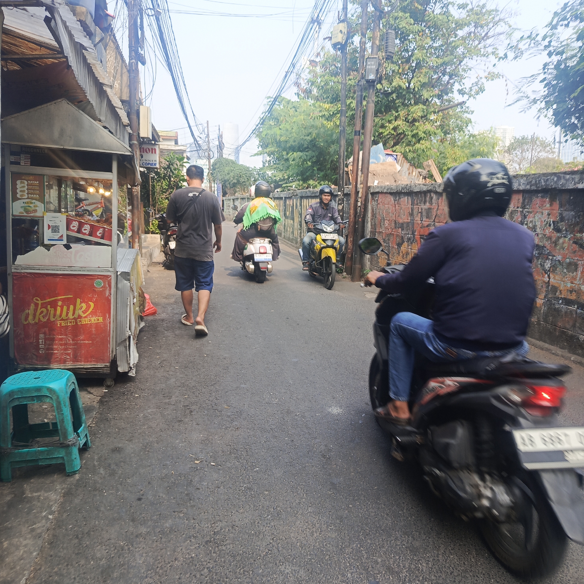 A man in a dark t-shirt and blue shorts walks away from the camera down a narrow, paved street. To his left is a food cart selling "Dkruk Fried Chicken". In front of the cart is a teal plastic stool. Several motorcycles are on the street. One motorcycle with a yellow body and a rider in a dark jacket and blue jeans is approaching the camera. Another motorcycle, a white scooter with a woman in a green headscarf riding on the back, is ahead of the yellow motorcycle, also approaching. Further ahead, another motorcycle is visible from behind, with a rider wearing a dark jacket and helmet. The street is lined with buildings and a brick wall on the right. Trees and power lines are visible above. The sky is overcast.