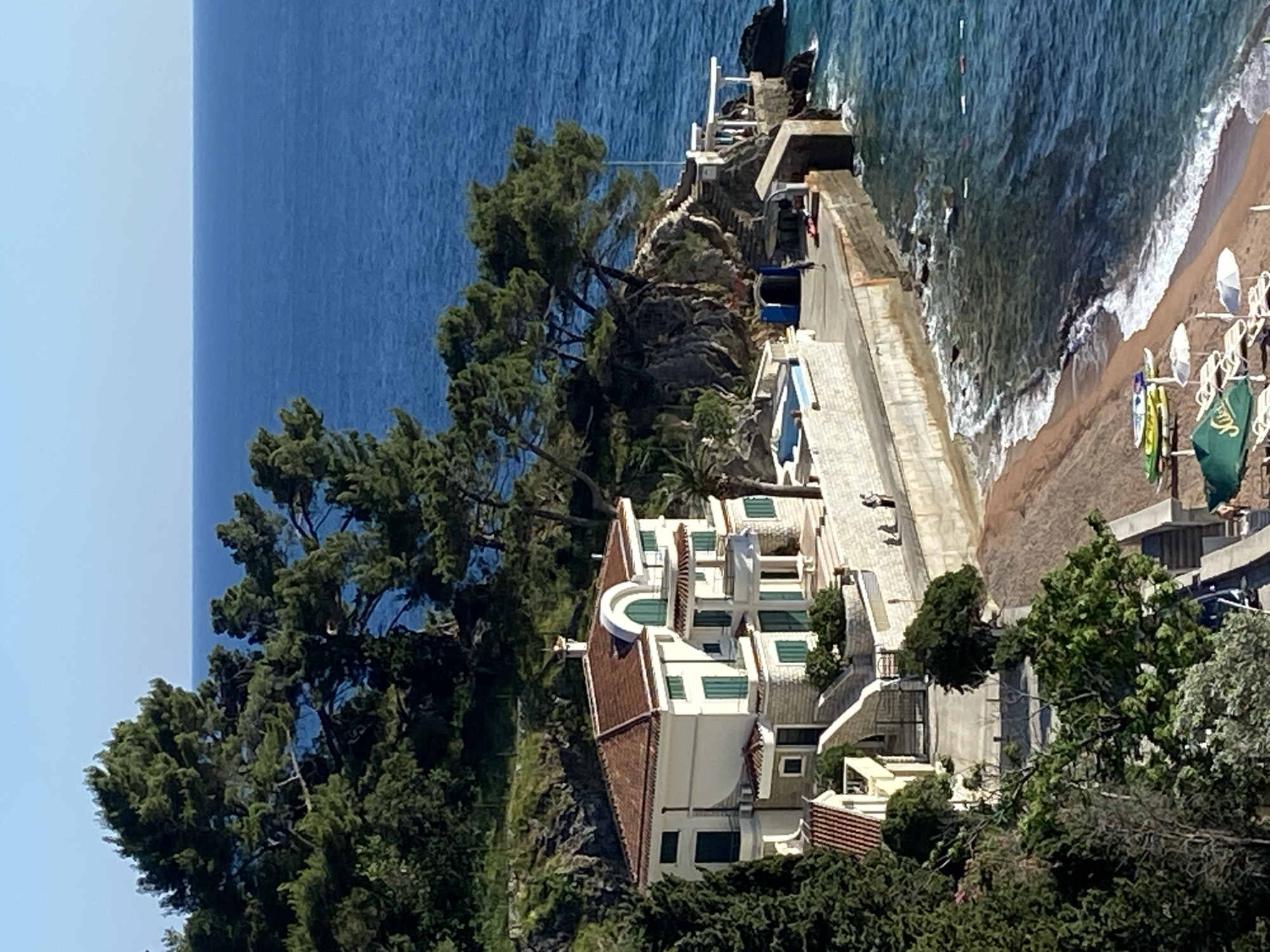 The image captures a coastal scene in Petrovac na Moru, Montenegro. A large, multi-story white building with a red tile roof sits on a cliff overlooking the Adriatic Sea. The building features multiple windows, green shutters, and a semi-circular balcony. Lush, green trees and vegetation surround the building. A concrete promenade runs along the edge of the cliff, with steps leading down to a small, sandy beach where people are sunbathing and relaxing under umbrellas. The water is clear and blue. The sky is light blue.