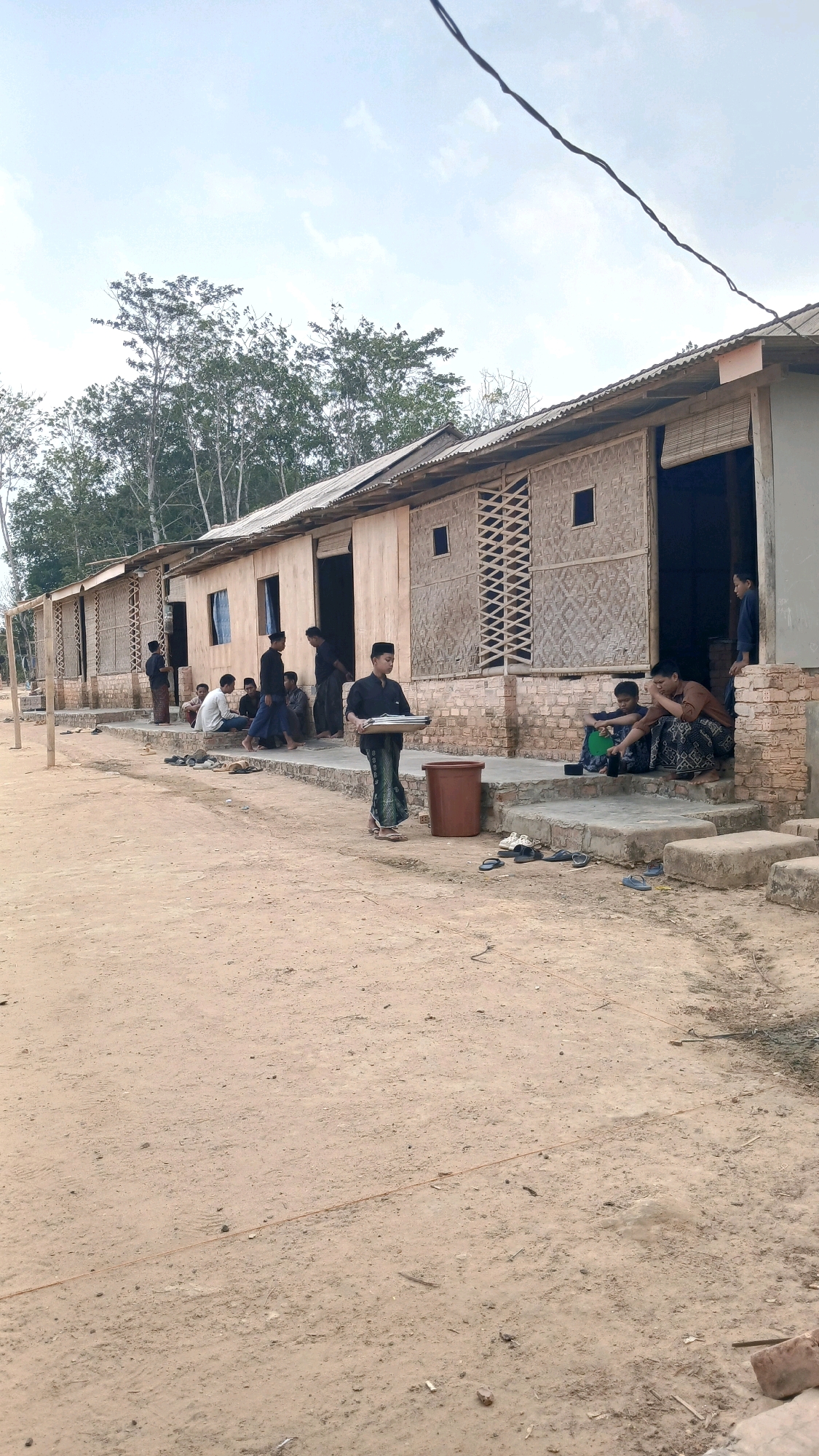 In Adi Luhur, Indonesia, a group of young men are gathered outside a row of simple, wooden buildings with corrugated metal roofs. The buildings appear to be single-story structures with woven bamboo panels on some walls and brick bases. Several men are seated on the steps leading into the buildings, while others stand nearby. One young man, wearing a dark shirt and patterned sarong, is walking forward carrying a flat tray with what appear to be food items. A brown trash bin is located on the steps. The ground is a dirt path. Trees are visible in the background, and the sky is overcast.