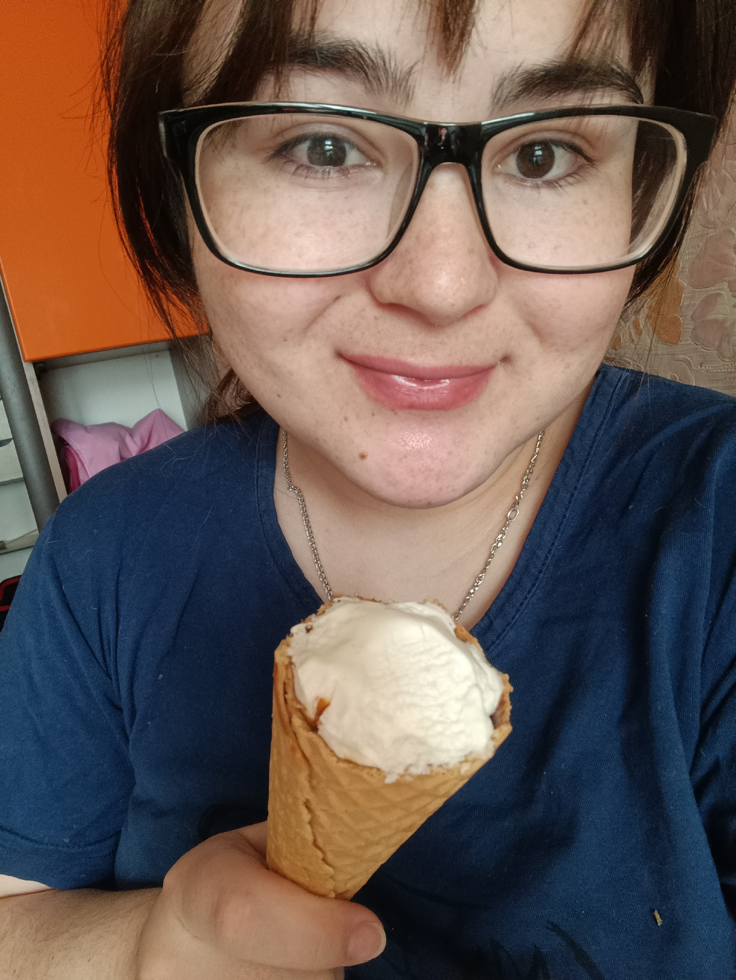 A woman with brown hair and glasses is holding an ice cream cone. She is wearing a blue t-shirt and a silver necklace. The background includes an orange cabinet. The location is likely not Noviy, Russia, as no identifying information is provided.