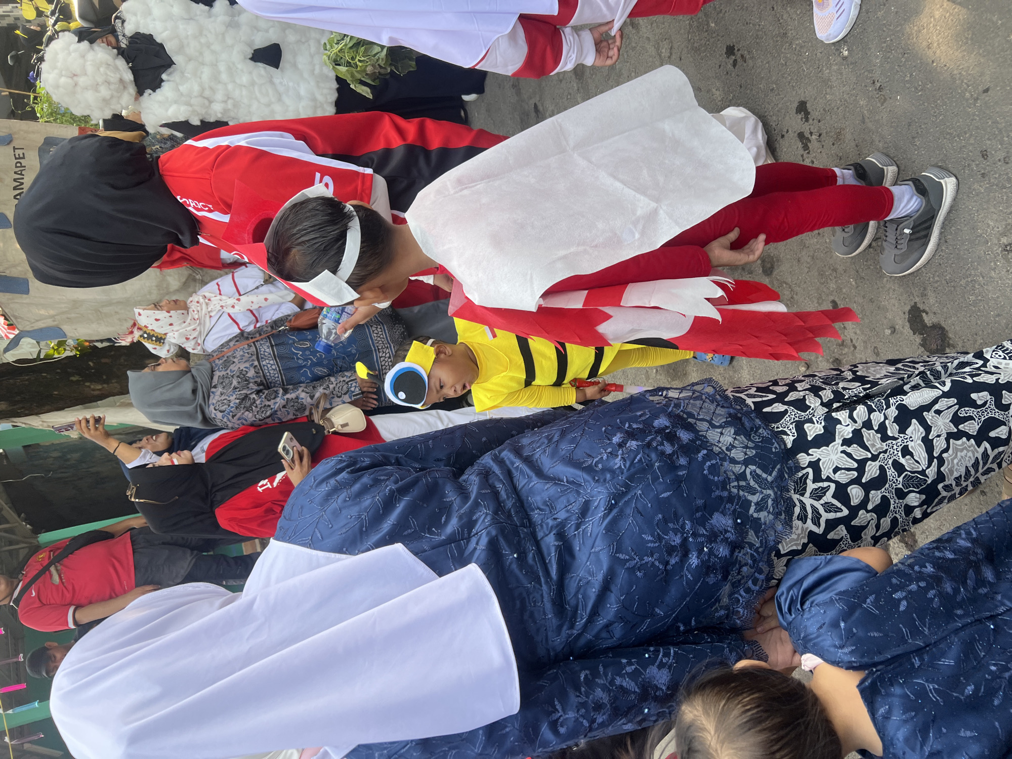 In Klapanunggal, Indonesia, a group of people, including children dressed in costumes, are gathered outdoors. The child in the foreground is wearing a bee costume with a yellow and black striped jumpsuit and antennae on a headband. Another child is dressed in a red and white superhero-like costume with a white cape. Adults are visible wearing a variety of attire, including a woman in a blue batik dress and headscarf, and others in red shirts and headscarves. The environment appears to be a street or open area, possibly for a local event or parade, with some decorative elements and foliage visible in the background. The overall scene suggests a festive community gathering.